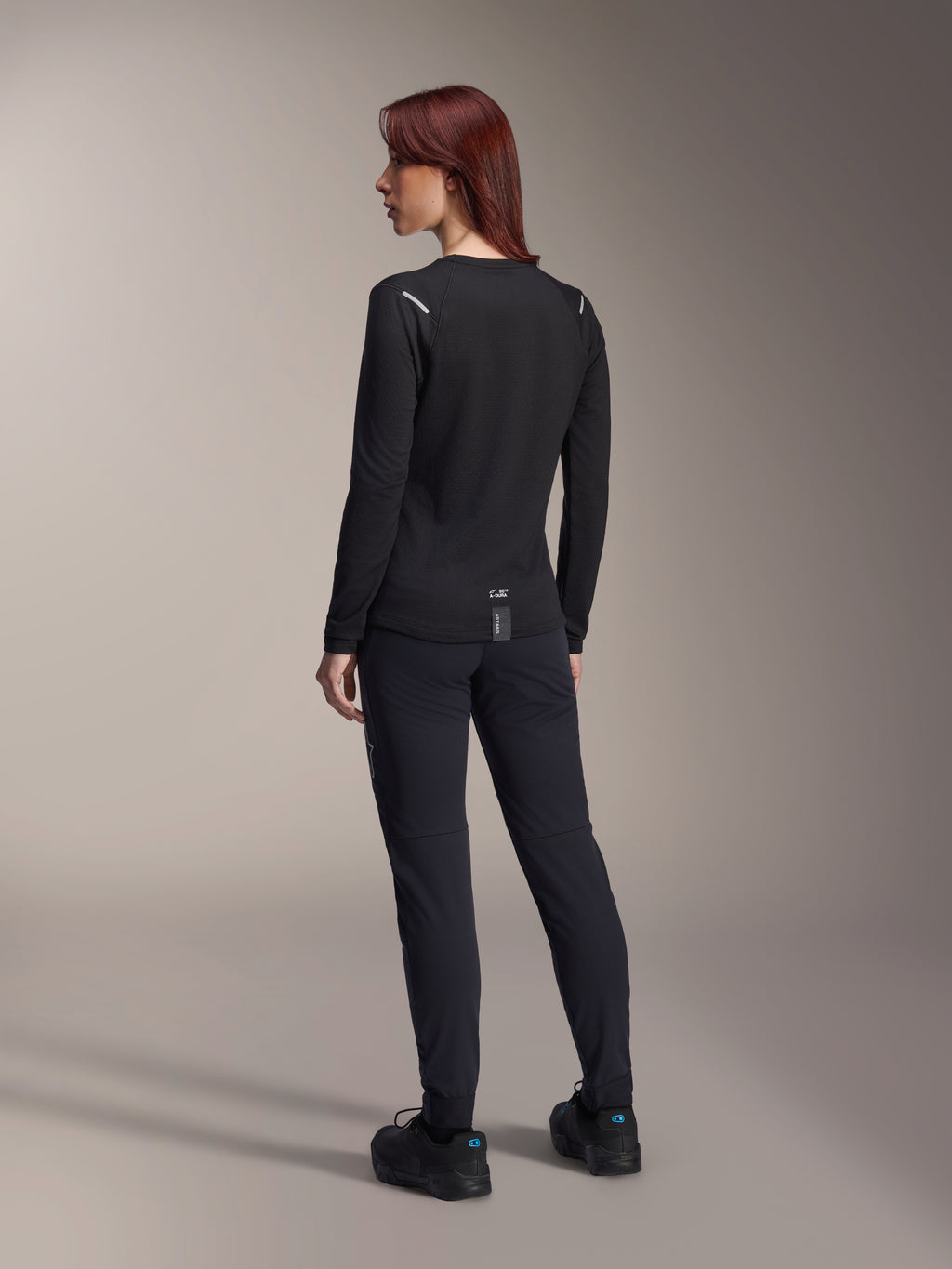 Alpinestars Stella A-Dura Thermal Jersey da donna, maglia da MTB a maniche lunghe in nero, vista posteriore con dettagli riflettenti sulle spalle e coda allungata, indossata con pantaloni e scarpe neri, abbigliamento tecnico per mountain bike