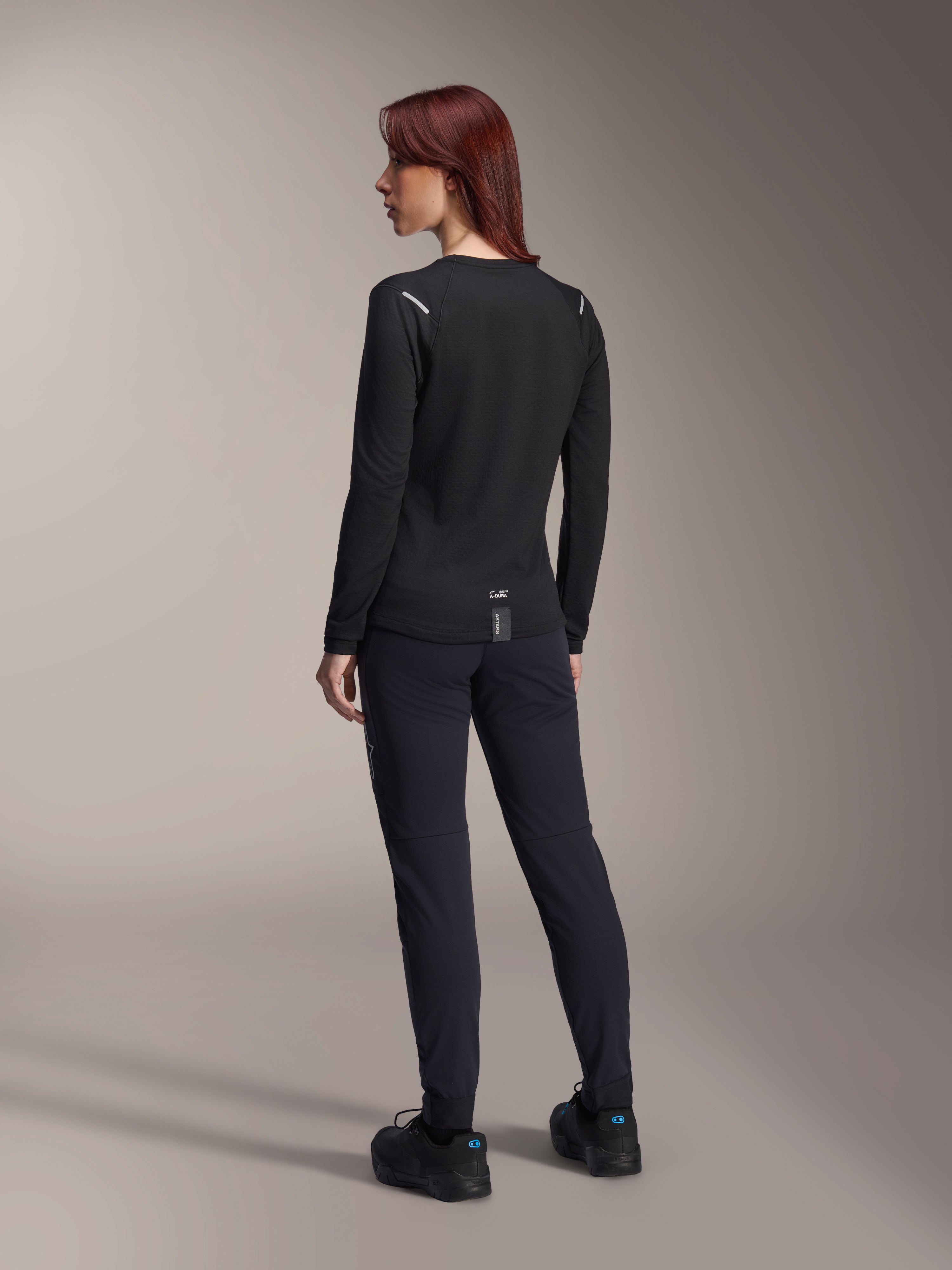 Alpinestars Stella A-Dura Thermal Jersey da donna, maglia da MTB a maniche lunghe in nero, vista posteriore con dettagli riflettenti sulle spalle e coda allungata, indossata con pantaloni e scarpe neri, abbigliamento tecnico per mountain bike