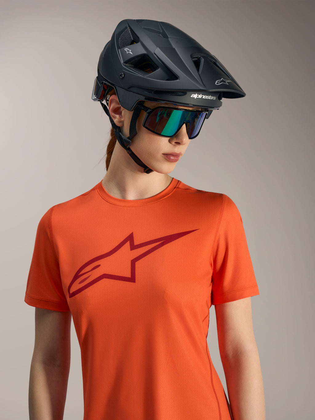 Maglia Alpinestars Stella A-Dura Ride arancione a maniche corte, logo rosso, casco MTB nero e occhiali iridescenti.