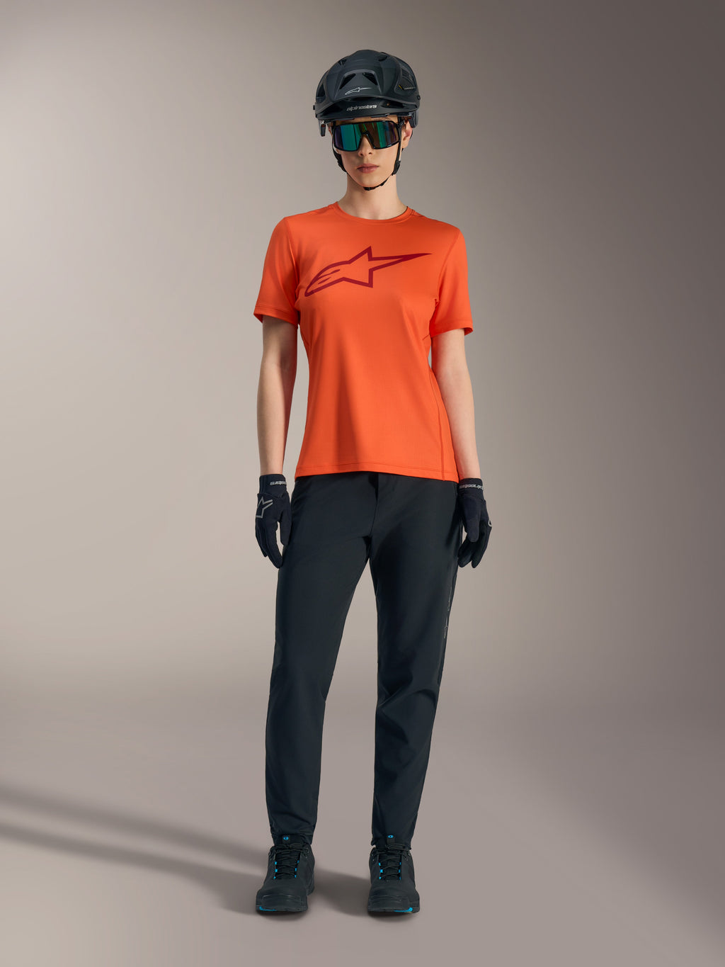 Maglia Alpinestars Stella A-Dura Ride arancione con logo bordeaux, abbinata a outfit MTB nero, casco e guanti.