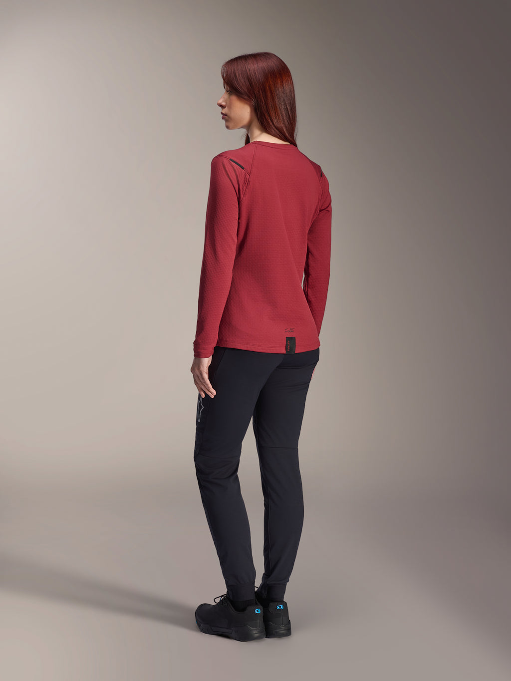 Donna che indossa i pantaloni termici Alpinestars Stella A-Dura neri, abbinati a una maglia termica rossa a maniche lunghe e scarpe nere, vista posteriore, abbigliamento da mountain bike per climi freddi