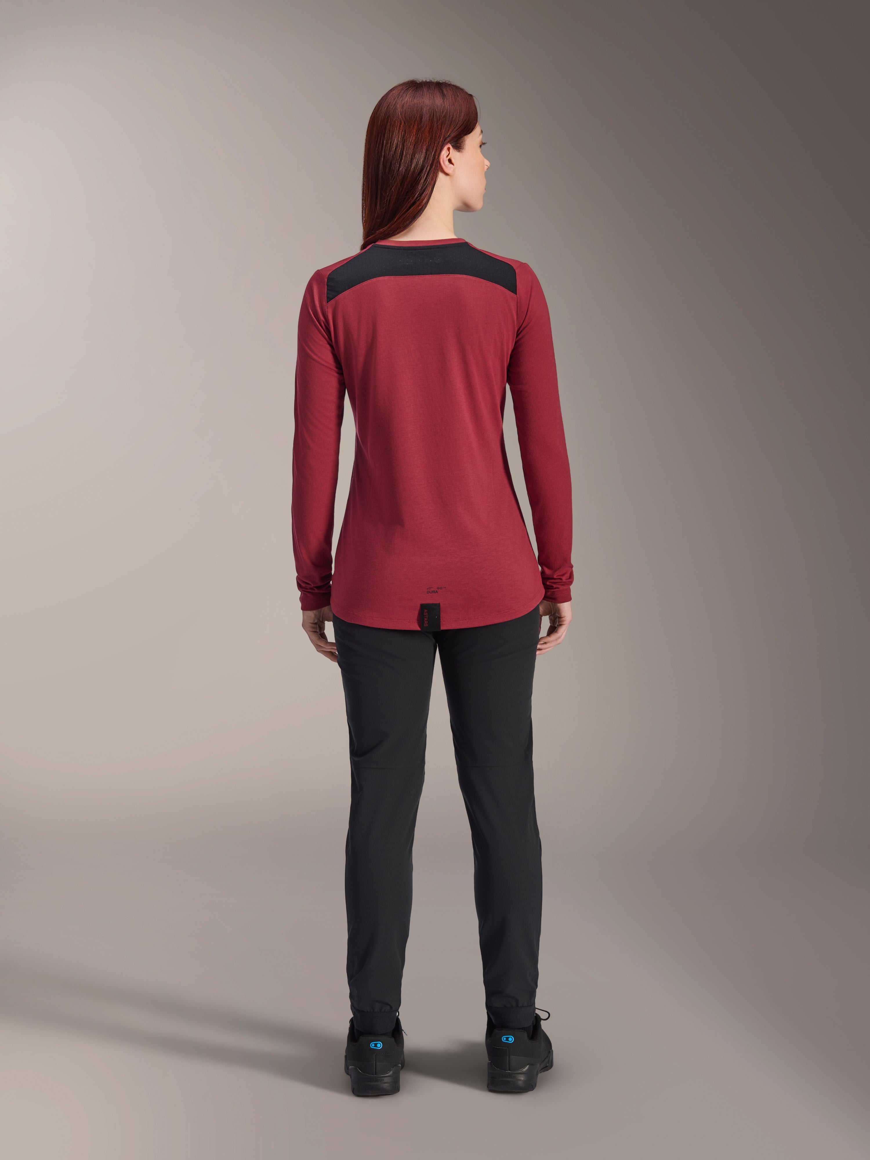 Maglia Alpinestars Stella A-Dura Dri Astar Long Sleeve da donna, rosso borgogna con pannelli in mesh nero sulle spalle, abbigliamento tecnico da MTB visto dal retro con pantaloni neri, design con coda lunga per maggiore copertura durante il ciclismo