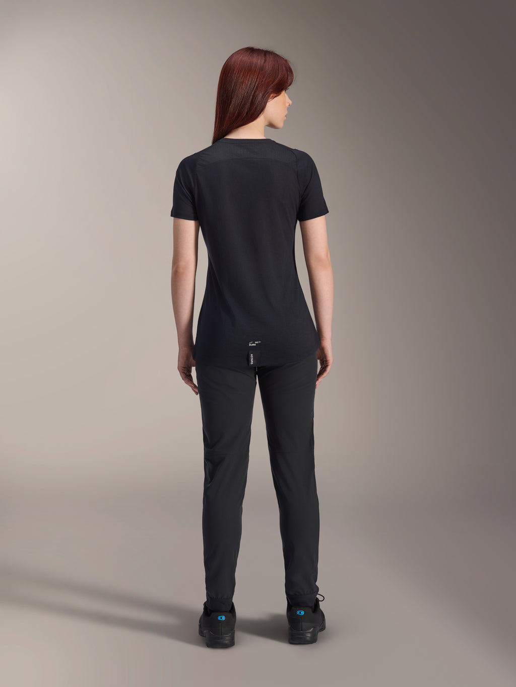 Alpinestars Stella A-Dura Dri Switch Short Sleeve Jersey da donna, nero, vista posteriore su una modella, con coda posteriore più lunga e pannelli in mesh tecnico, abbinata a pantaloni neri, abbigliamento tecnico da ciclismo MTB