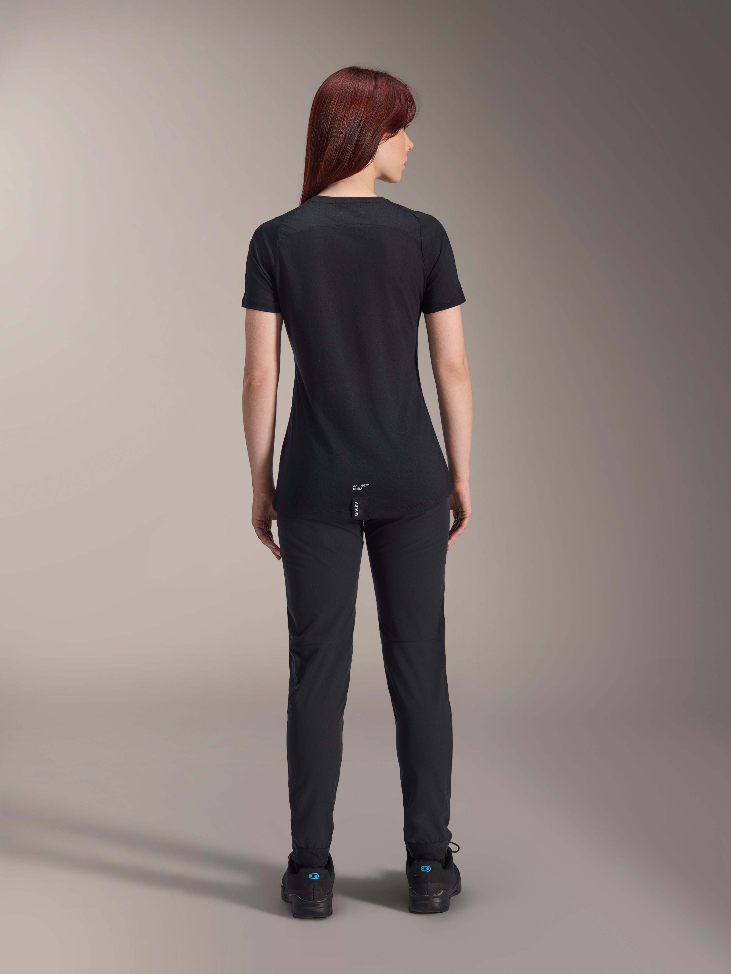Alpinestars Stella A-Dura Dri Switch Short Sleeve Jersey da donna, nero, vista posteriore su una modella, con coda posteriore più lunga e pannelli in mesh tecnico, abbinata a pantaloni neri, abbigliamento tecnico da ciclismo MTB