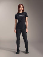 Modella con maglia a maniche corte Alpinestars Stella A-Dura Dri Switch da donna in nero, logo bianco sul petto, pantaloni tecnici neri e scarpe sportive nere, scatto in studio a figura intera