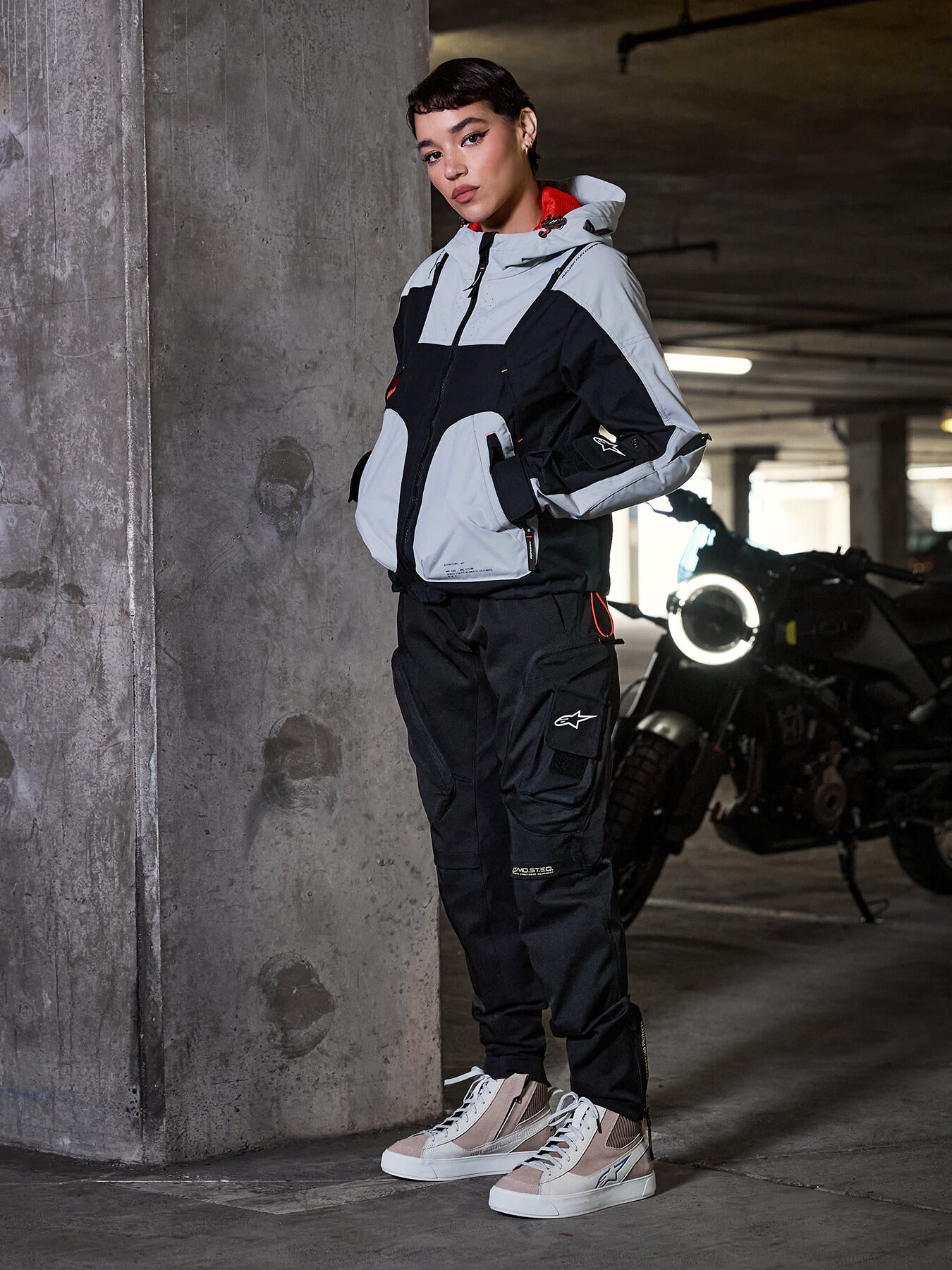 Pantaloni da donna Alpinestars Actea, neri, motociclista che indossa pantaloni da moto in stile cargo con una giacca tecnica grigia e nera e sneakers beige, ambientazione in un parcheggio urbano con motocicletta.