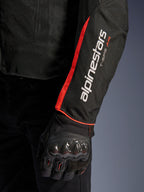 Giacca Alpinestars T-Sps Air V2, primo piano della manica in Black Red Fluo, con loghi Alpinestars e T-SPS AIR bianchi e striscia rosso fluorescente brillante, indossata con un guanto protettivo da moto nero con protezione delle nocche in fibra di carbonio