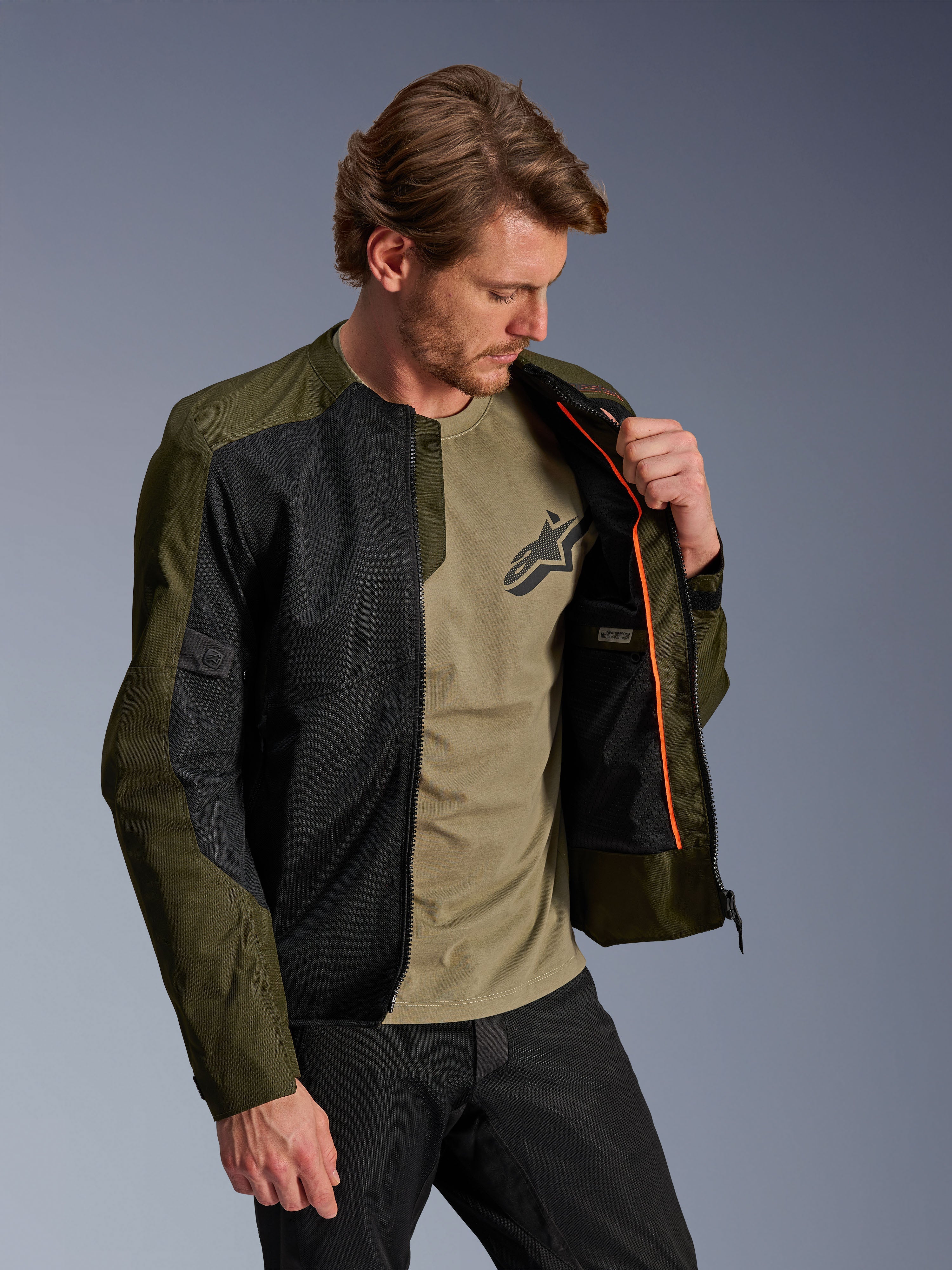 Giacca Alpinestars C-1 Air in Khaki e Nero, indossata da un modello per mostrare la fodera interna arancione e lo scomparto impermeabile, caratterizzata da ampi pannelli in rete tecnica per la ventilazione, stile urban contemporaneo abbinato a una t-shirt oliva e pantaloni neri