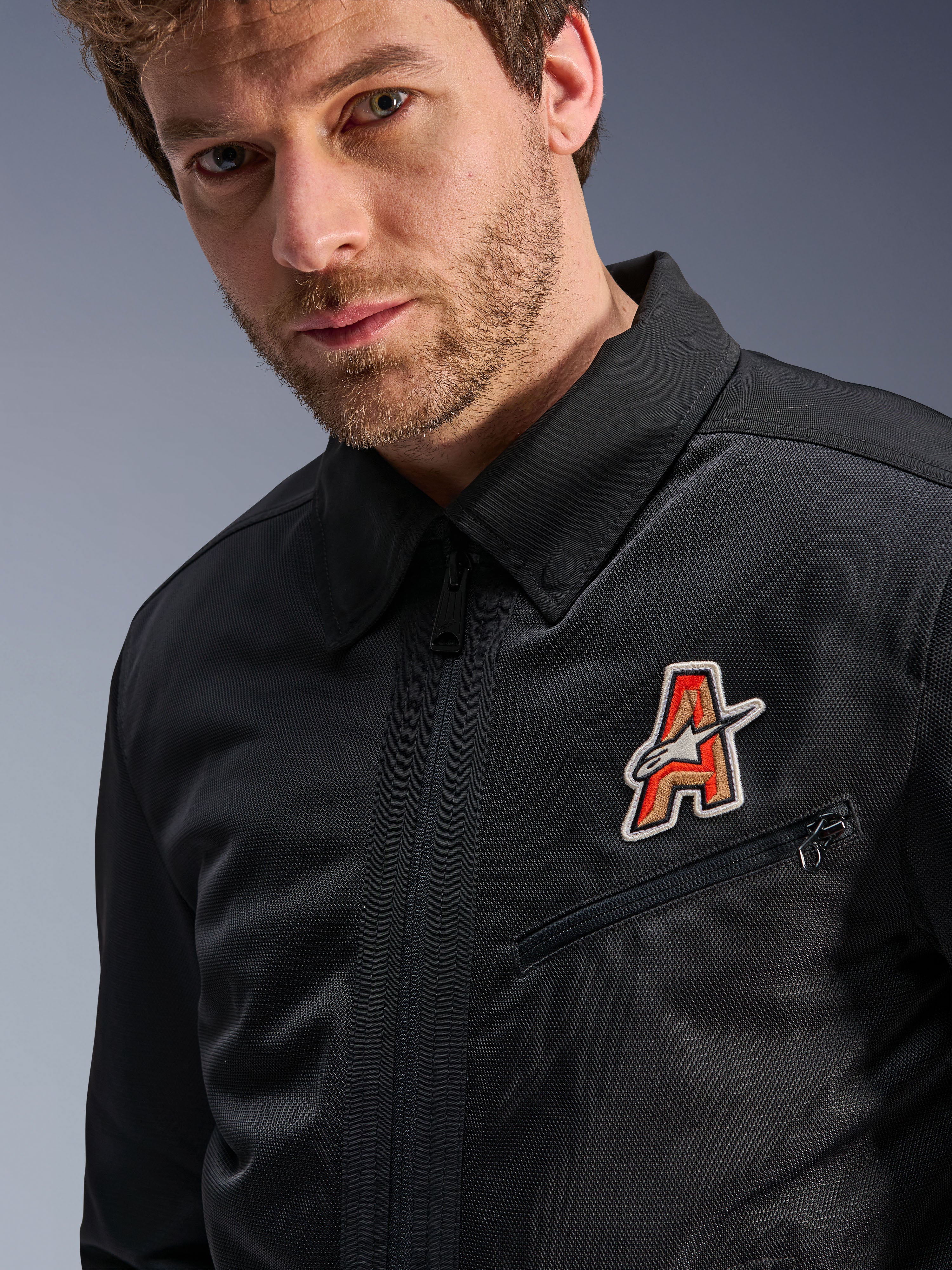 Giacca Alpinestars Ignite Air, nera, con colletto a camicia tradizionale e struttura in mesh traspirante, caratterizzata da una toppa con logo 'A' rosso e oro sul petto, indossata da un modello uomo, abbigliamento moto urbano