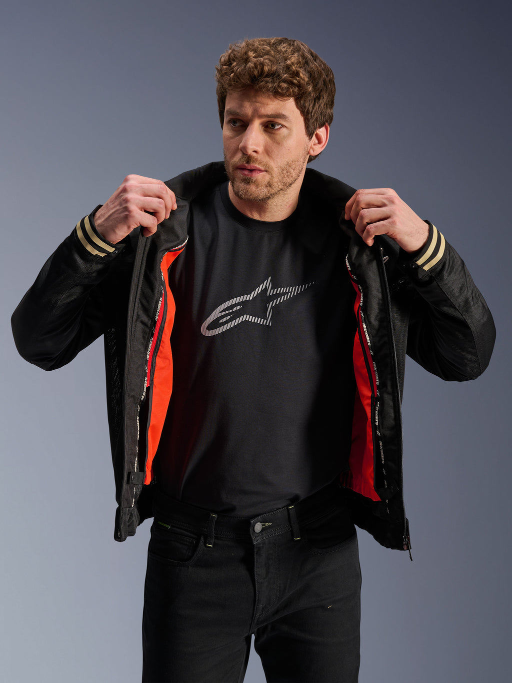 Uomo con giacca Alpinestars Ignite Air, nera, caratterizzata da corpo in rete e fodera antivento staccabile arancione brillante, abbinata a t-shirt nera con logo Astars e pantaloni neri, stile motociclistico urbano contemporaneo