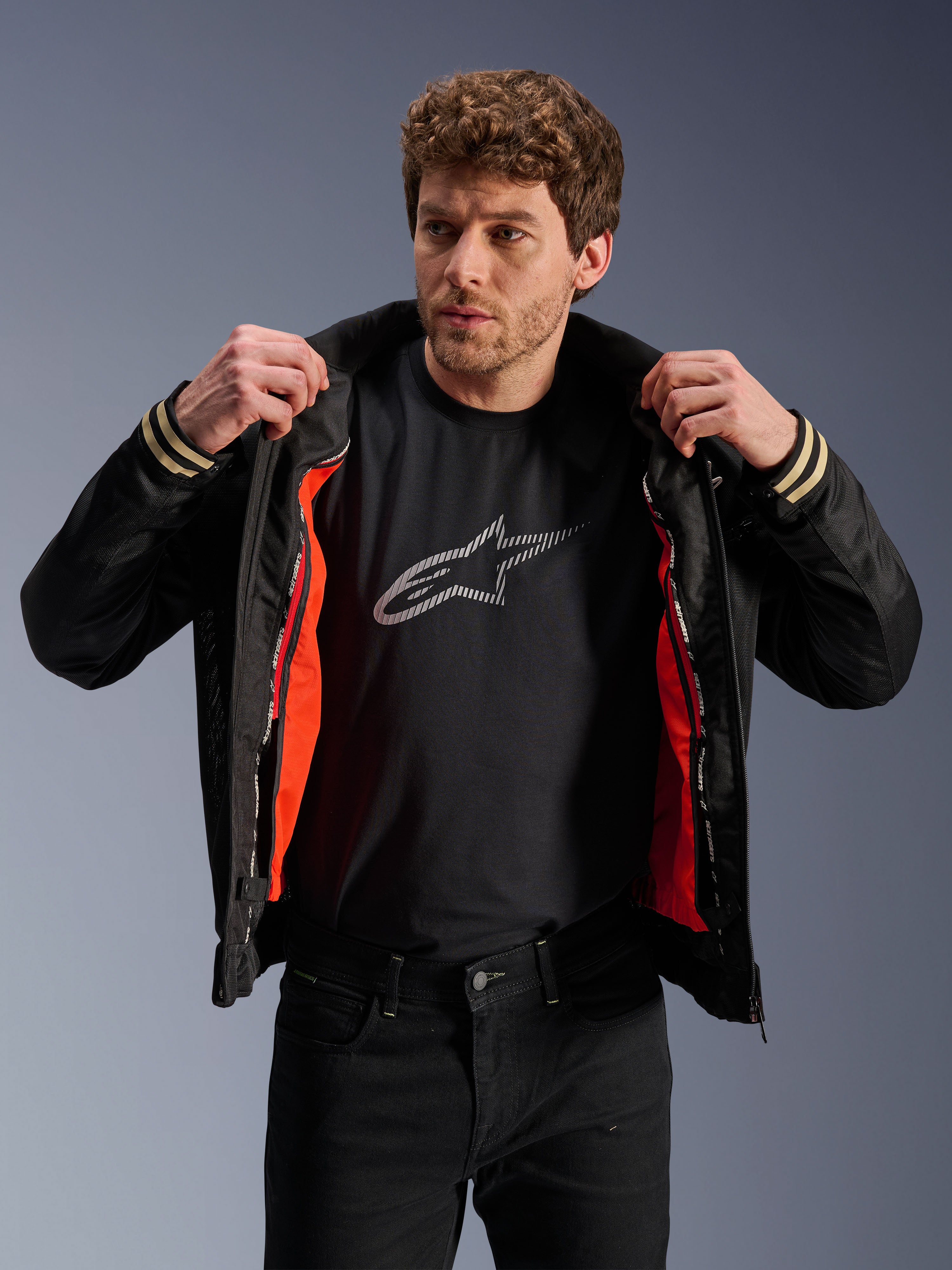 Uomo con giacca Alpinestars Ignite Air, nera, caratterizzata da corpo in rete e fodera antivento staccabile arancione brillante, abbinata a t-shirt nera con logo Astars e pantaloni neri, stile motociclistico urbano contemporaneo