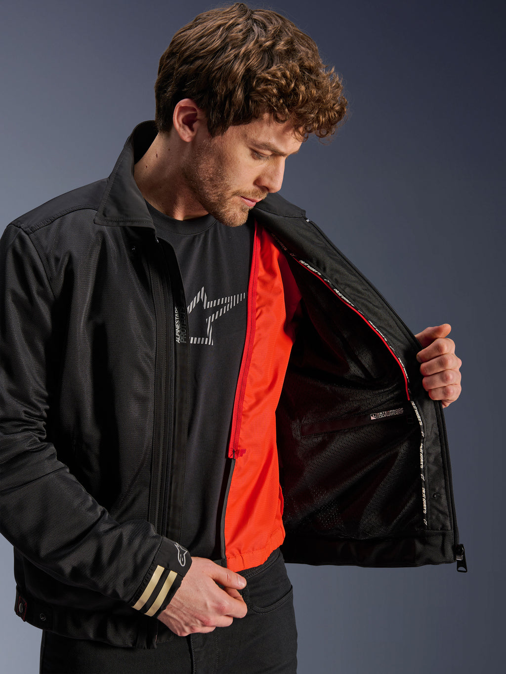 Alpinestars Ignite Air Jacket, giacca da moto urban nera, con fodera antivento staccabile arancione brillante e corpo in rete, vista interna dello scomparto impermeabile, indossata con t-shirt nera con logo Alpinestars