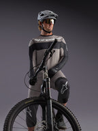 Alpinestars A-Dura Race, maglia grigia a maniche lunghe su rider MTB, dettagli neri e abbigliamento tecnico professionale.