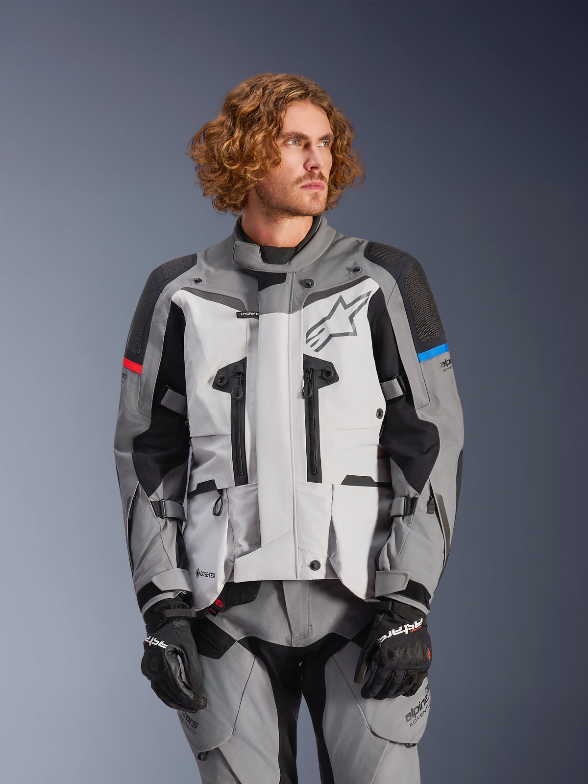 Alpinestars Boulder Gore-Tex, giacca da moto touring, grigia e nera con dettagli rossi e blu, impermeabile e traspirante, indossata con pantaloni coordinati e guanti neri