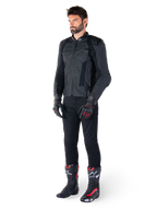 Giacca in pelle Alpinestars Missile V3, nero, indossata da un pilota con pantaloni neri e guanti e stivali da corsa neri e rossi, con pannelli in pelle traforata e branding monocromatico, estetica racing professionale
