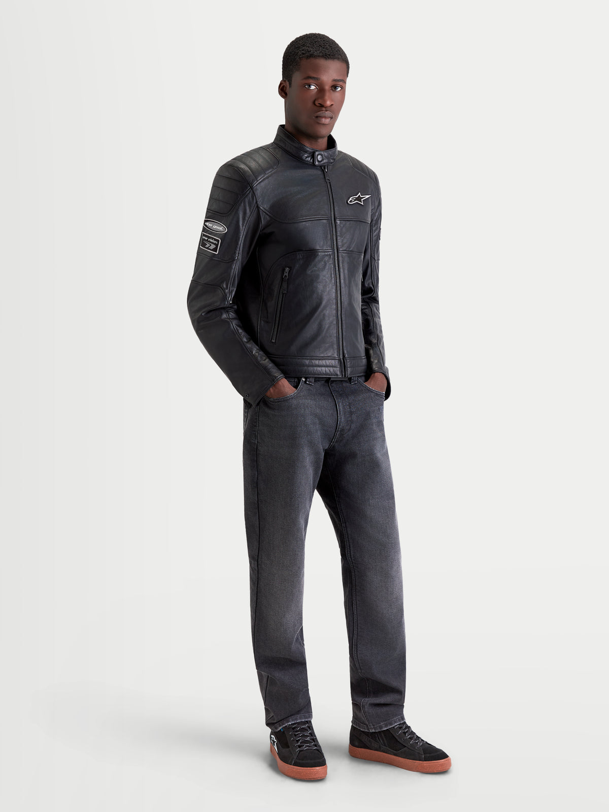 Alpinestars Revern Giacca di pelle, giacca da moto urbana in pelle da uomo, nero, vista frontale su un modello che indossa jeans scuri e scarpe da moto, con dettagli in stile heritage e struttura in pelle bovina pieno fiore effetto vintage