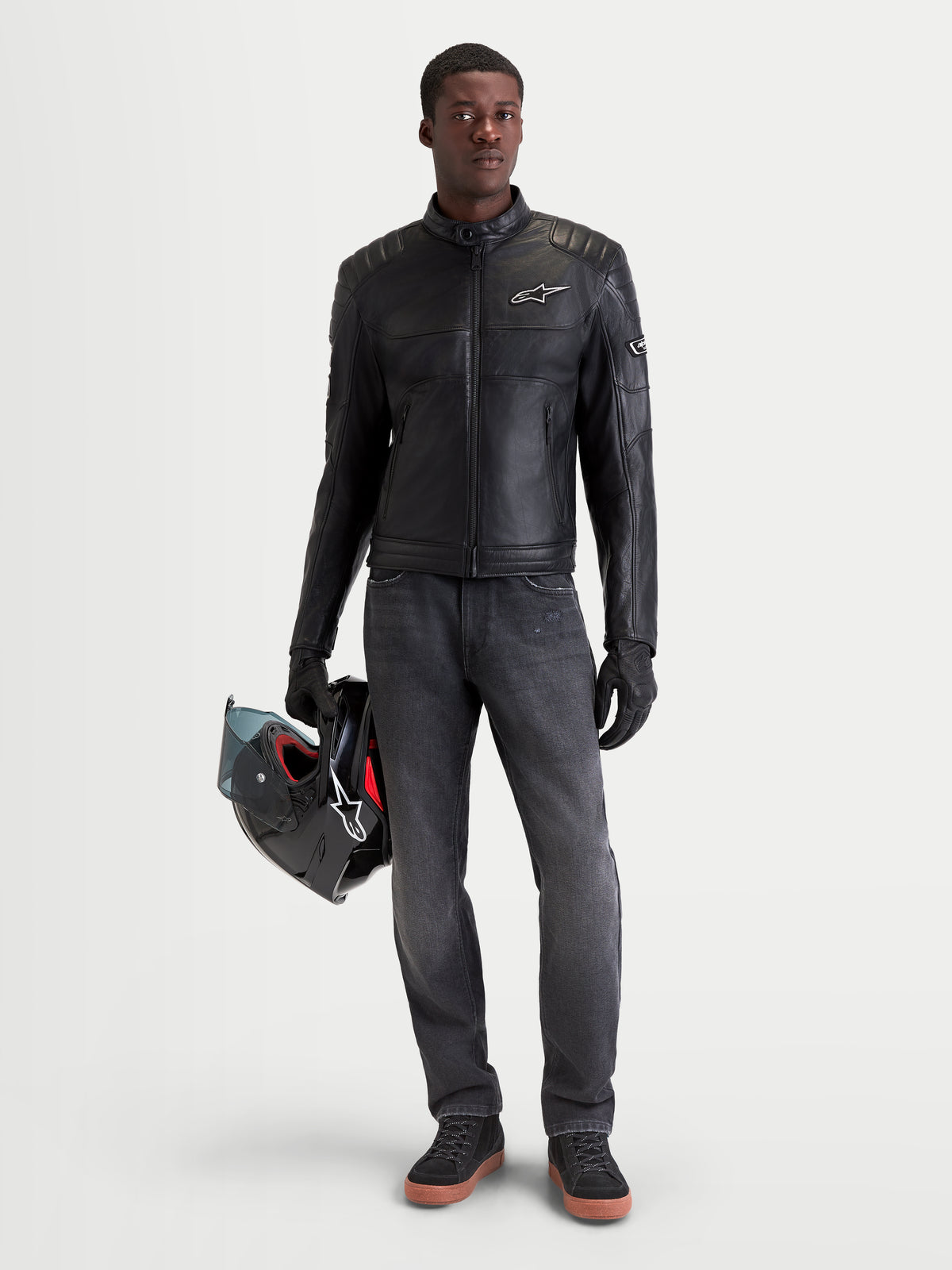 Alpinestars Revern Giacca di pelle, giacca da moto urbana da uomo in pelle nera, vista frontale su un modello che indossa jeans scuri e tiene un casco