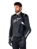 Uomo che indossa la giacca in pelle Alpinestars Missile V3 Airflow, giacca da moto in nero e bianco, pelle pregiata traforata per il flusso d'aria, logo Alpinestars bianco su petto e manica, vestibilità sportiva ispirata alle corse, indossata con guanti e pantaloni neri.