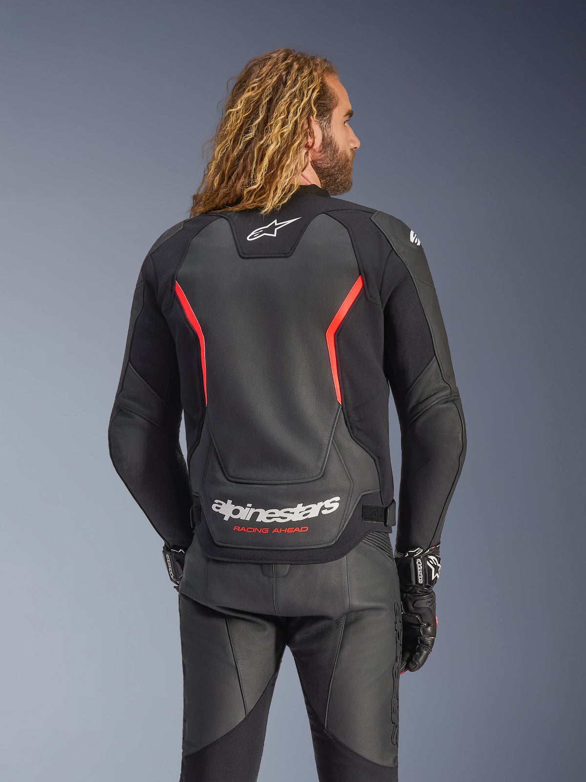 Giacca in pelle Alpinestars GP Force V2, nero e rosso fluo, vista posteriore su un modello, con logo Alpinestars bianco e scritta 'RACING AHEAD', abbinata a pantaloni e guanti da moto in pelle nera.