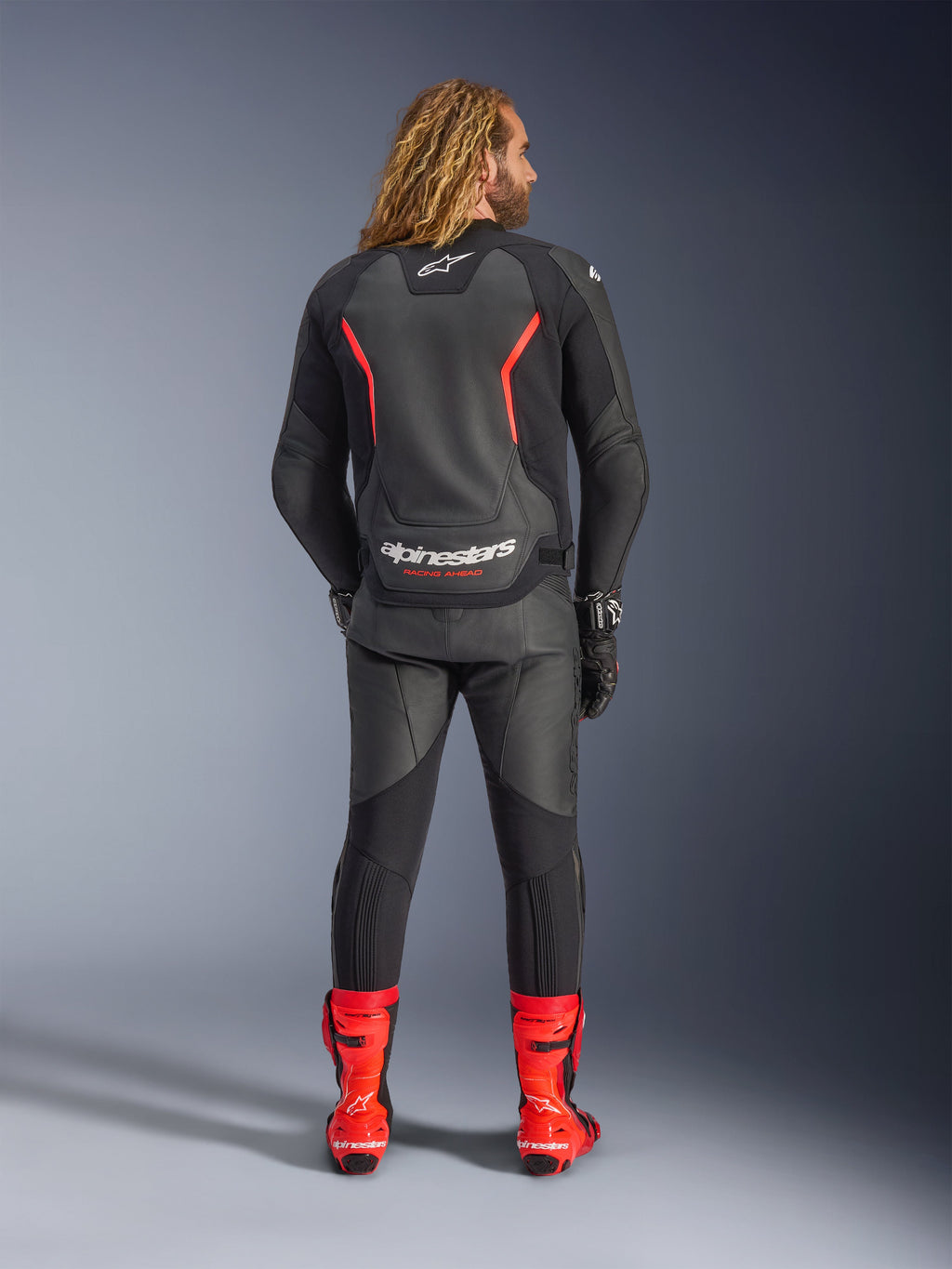Giacca in pelle Alpinestars GP Force V2, nero e rosso fluo, vista da dietro, indossata con pantaloni in pelle nera coordinati e stivali da corsa rosso acceso, con linee d'accento rosse e il marchio Alpinestars bianco sul retro, completo professionale da motociclismo sportivo.