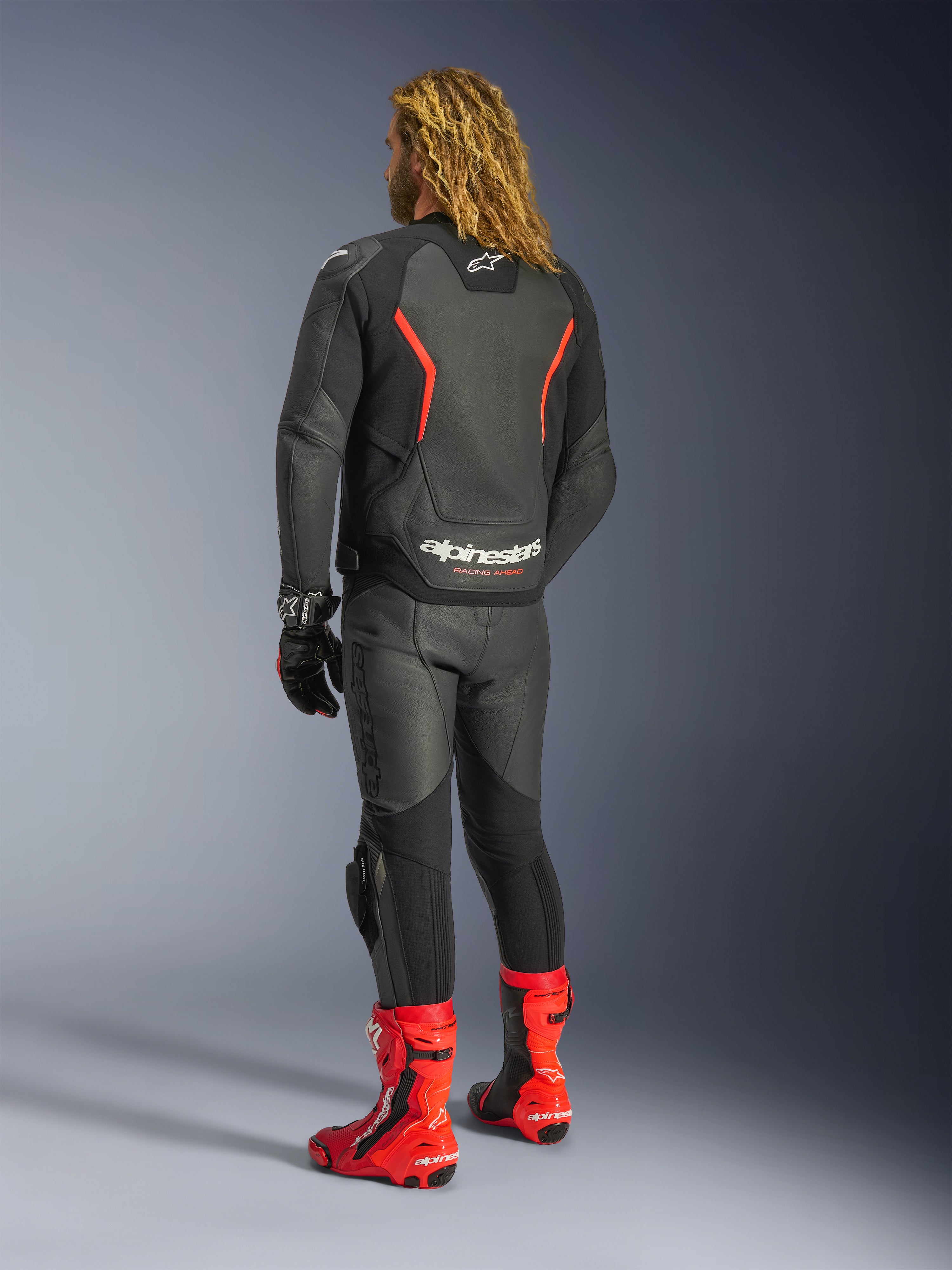 Alpinestars GP Force V2 Leather Jacket, Nero Rosso Fluo, vista posteriore indossata da un pilota con pantaloni in pelle nera coordinati, stivali racing rossi e guanti neri, con vivaci accenti rossi e logo Alpinestars sul retro, abbigliamento da moto con vestibilità sportiva