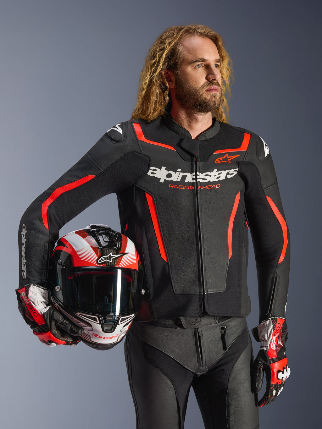Giacca in pelle Alpinestars GP Force V2, nero e rosso fluo, indossata da un modello che tiene un casco abbinato, con loghi Alpinestars bianchi e accenti rossi, abbinata a pantaloni in pelle e guanti da corsa, abbigliamento sportivo da moto per strada e pista.