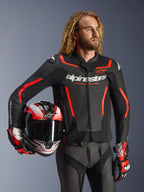 Giacca in pelle Alpinestars GP Force V2, nero e rosso fluo, indossata da un modello che tiene un casco abbinato, con loghi Alpinestars bianchi e accenti rossi, abbinata a pantaloni in pelle e guanti da corsa, abbigliamento sportivo da moto per strada e pista.
