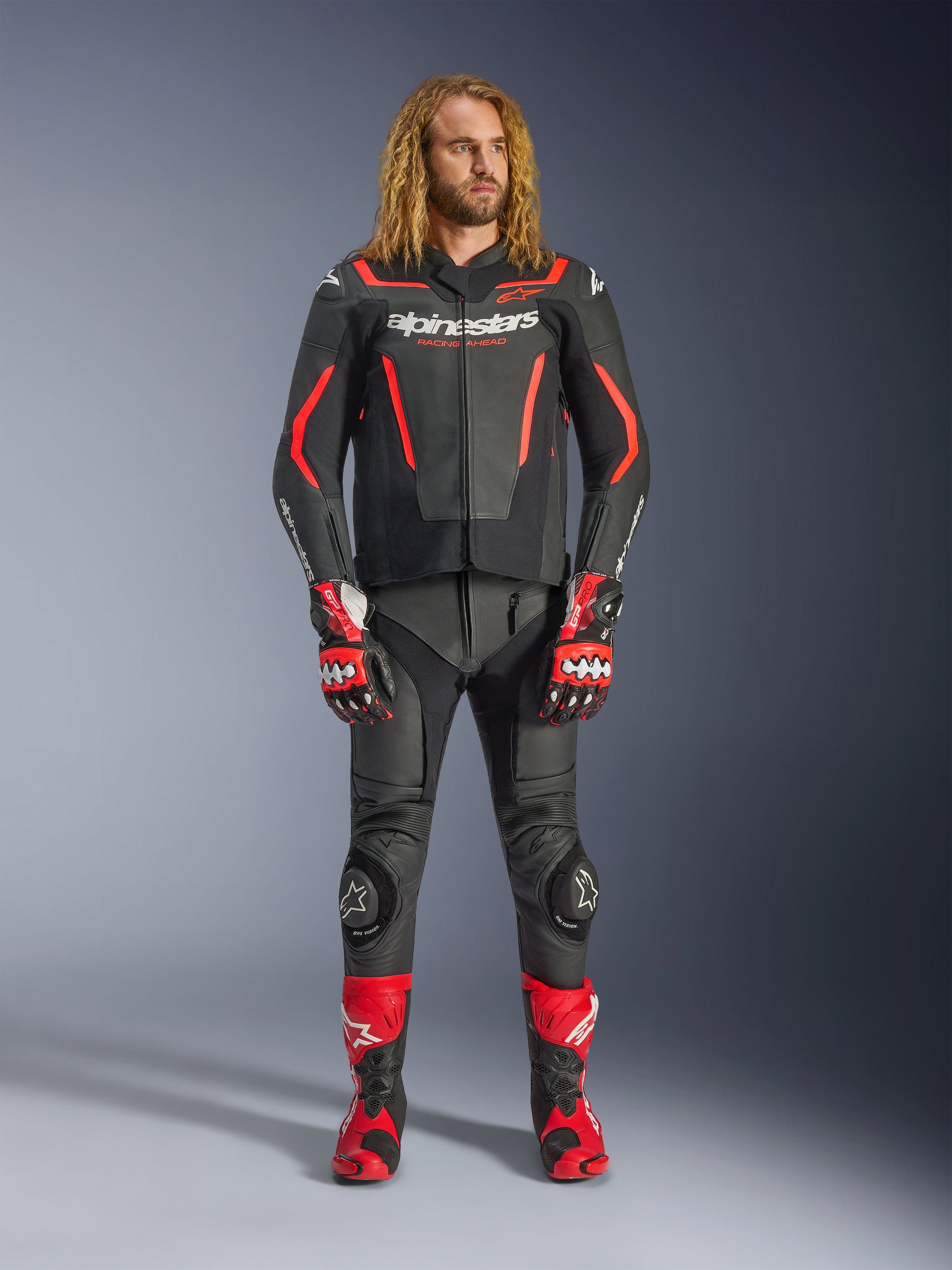 Modello uomo che indossa la giacca in pelle Alpinestars GP Force V2 in nero e rosso fluo, abbinata a pantaloni in pelle coordinati, guanti racing rossi e stivali performance rossi, abbigliamento sportivo da moto professionale