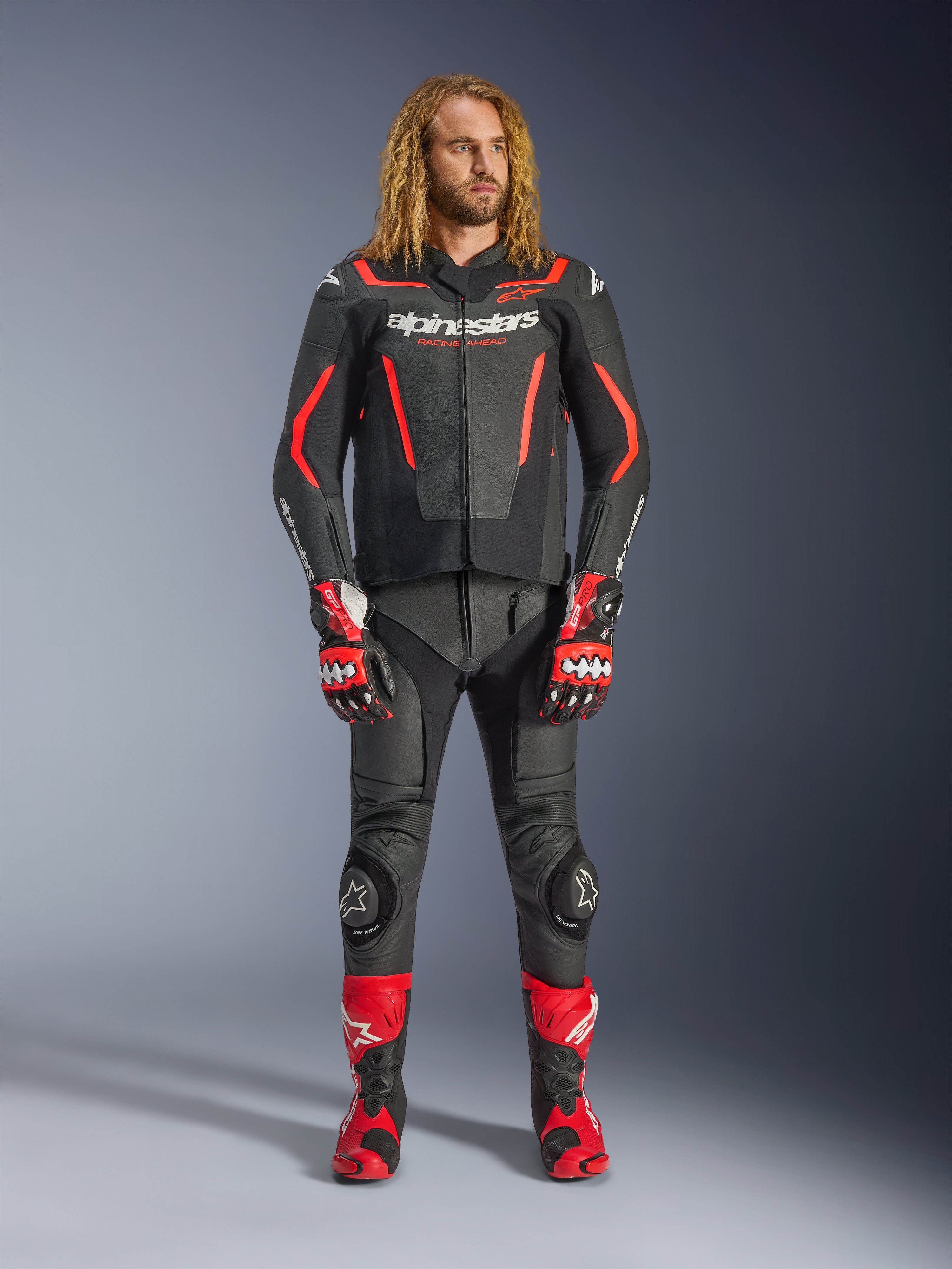 Modello uomo che indossa la giacca in pelle Alpinestars GP Force V2 in nero e rosso fluo, abbinata a pantaloni in pelle coordinati, guanti racing rossi e stivali performance rossi, abbigliamento sportivo da moto professionale