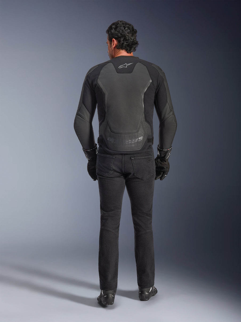 Giacca in pelle Alpinestars GP Force V2, nero, vista posteriore, giacca da moto sportiva premium con costruzione in pelle e tessuto, indossata con jeans da moto e guanti neri, con pannelli protettivi sulle spalle e dettagli del brand