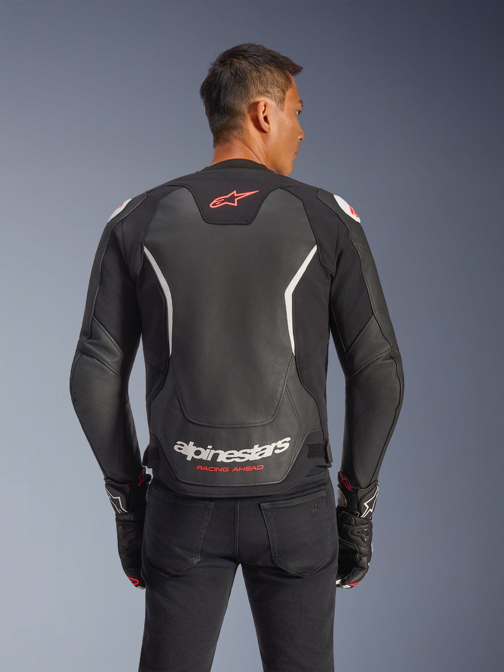 Giacca in pelle Alpinestars GP Force V2, vista posteriore, nero e bianco, con logo a stella rosso sulla parte superiore della schiena e marchio Alpinestars bianco con testo 'Racing Ahead', indossata con pantaloni neri e guanti coordinati, giacca da moto in pelle con vestibilità sportiva