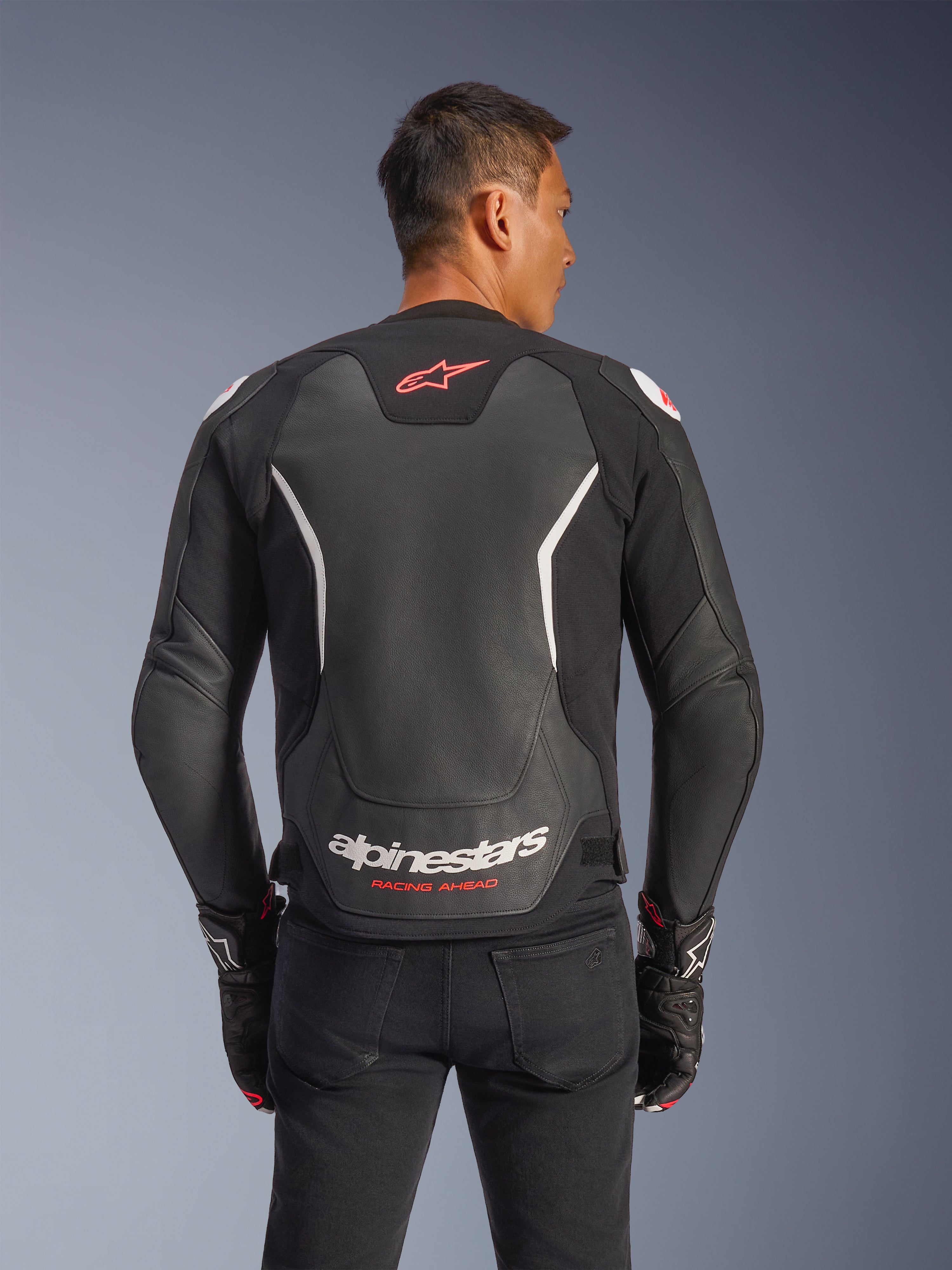 Giacca in pelle Alpinestars GP Force V2, vista posteriore, nero e bianco, con logo a stella rosso sulla parte superiore della schiena e marchio Alpinestars bianco con testo 'Racing Ahead', indossata con pantaloni neri e guanti coordinati, giacca da moto in pelle con vestibilità sportiva