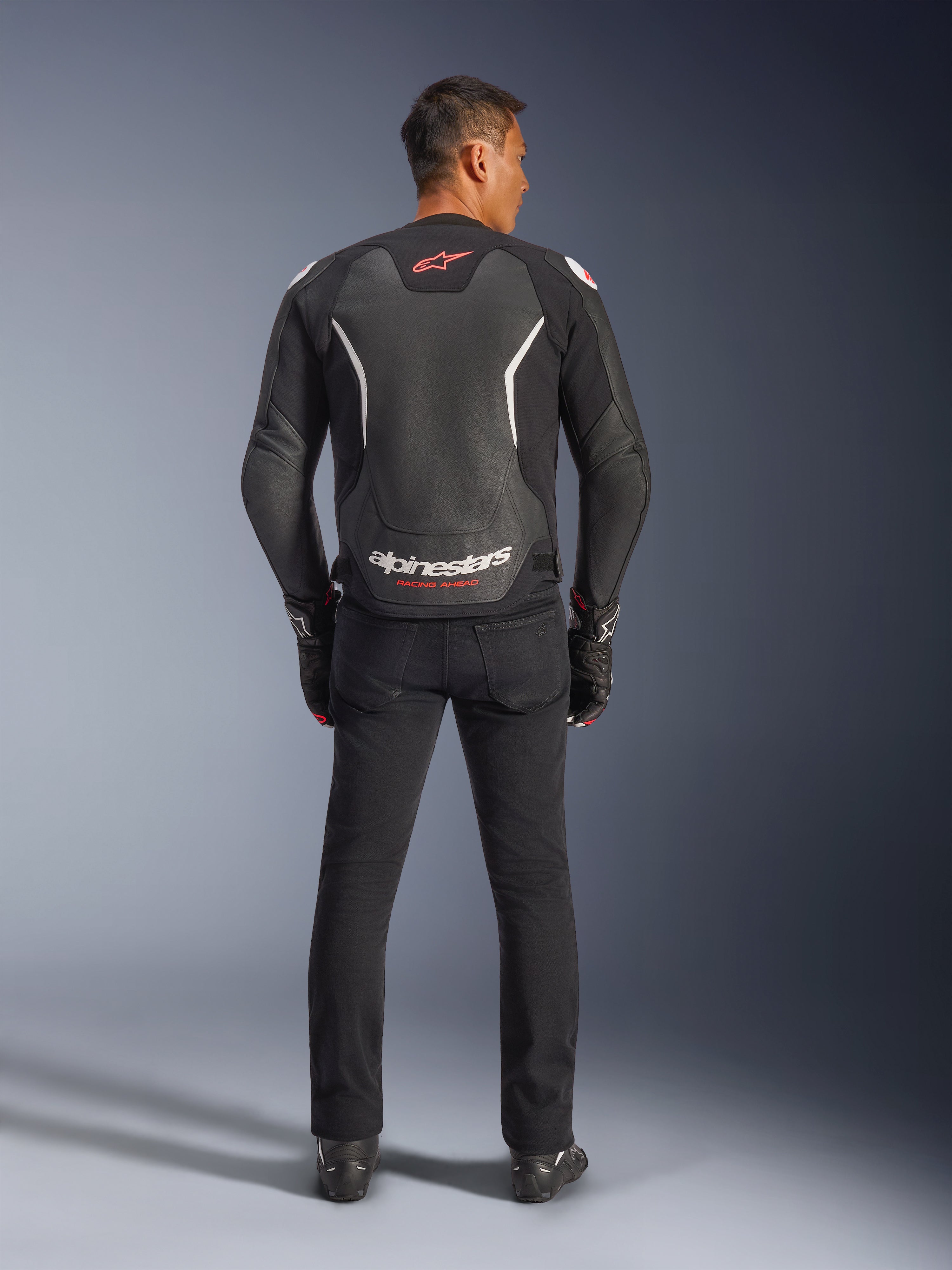 Giacca in pelle Alpinestars GP Force V2, nero e bianco, vista posteriore, indossata da un pilota con pantaloni e guanti neri, con grande logo bianco sulla parte bassa della schiena e dettagli rossi, realizzata in pregiata pelle bovina per abbigliamento moto stradale