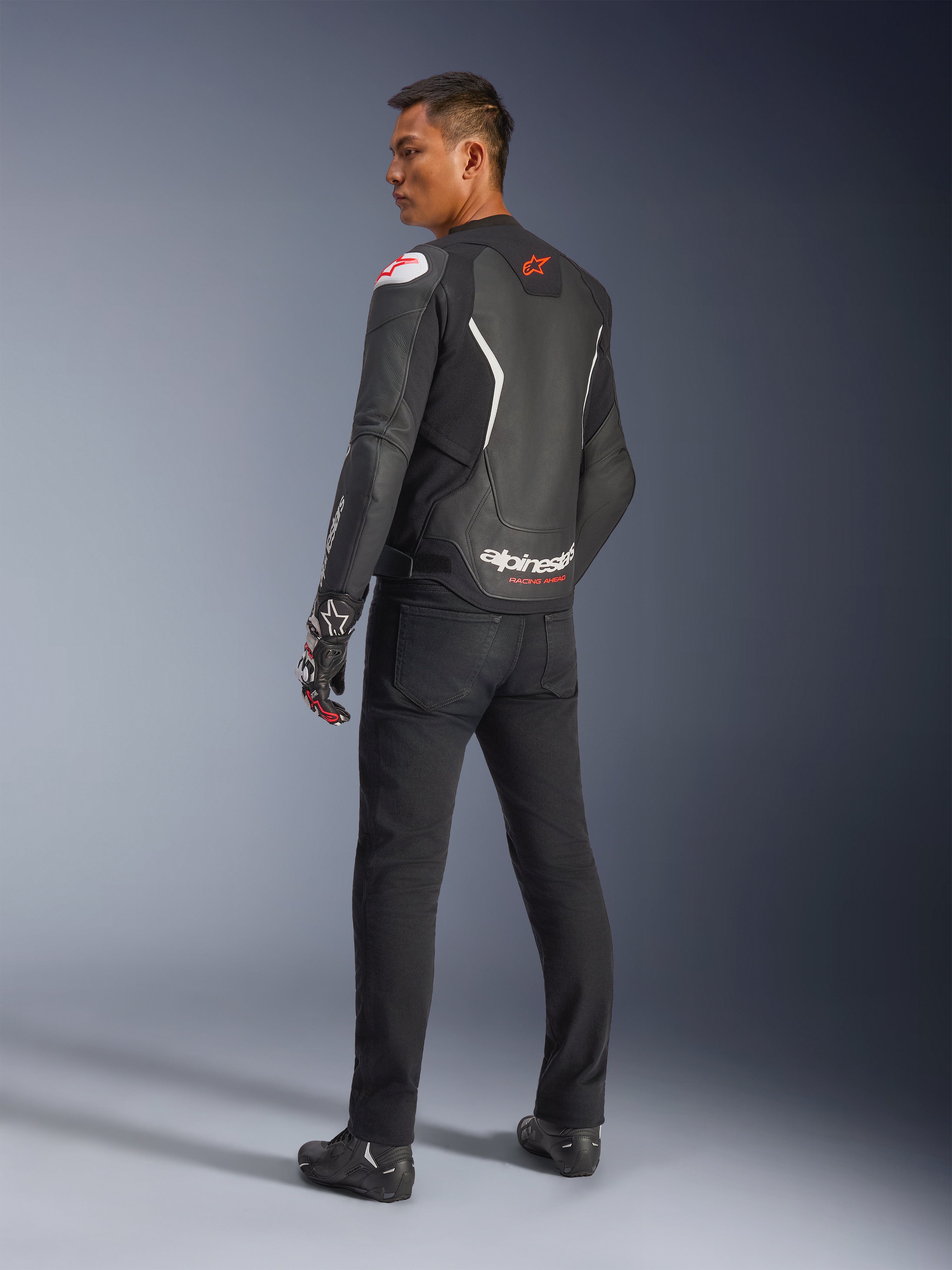Giacca in pelle Alpinestars GP Force V2, nero e bianco, vista posteriore che mostra la costruzione in pelle con accenti bianchi e loghi rossi, indossata con pantaloni neri e guanti racing, con evidente marchio Alpinestars sulla parte bassa della schiena, abbigliamento da moto sportivo