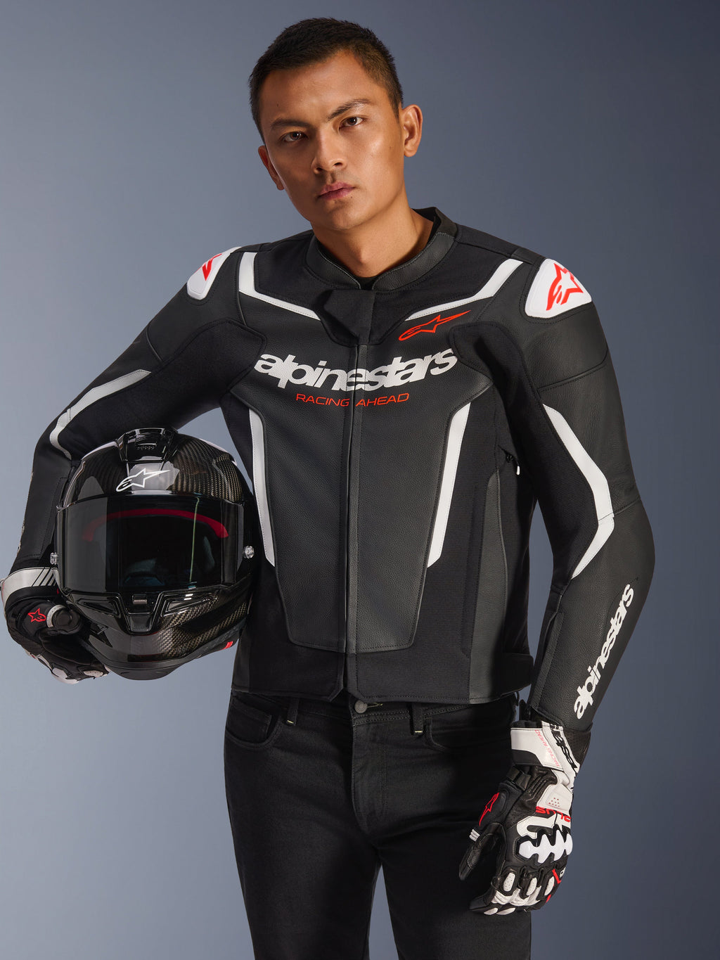 Giacca in pelle Alpinestars GP Force V2, nero e bianco con dettagli rossi, indossata da un modello che tiene un casco da moto in fibra di carbonio e indossa guanti da corsa, con pannelli bianchi a contrasto e marchio Alpinestars.