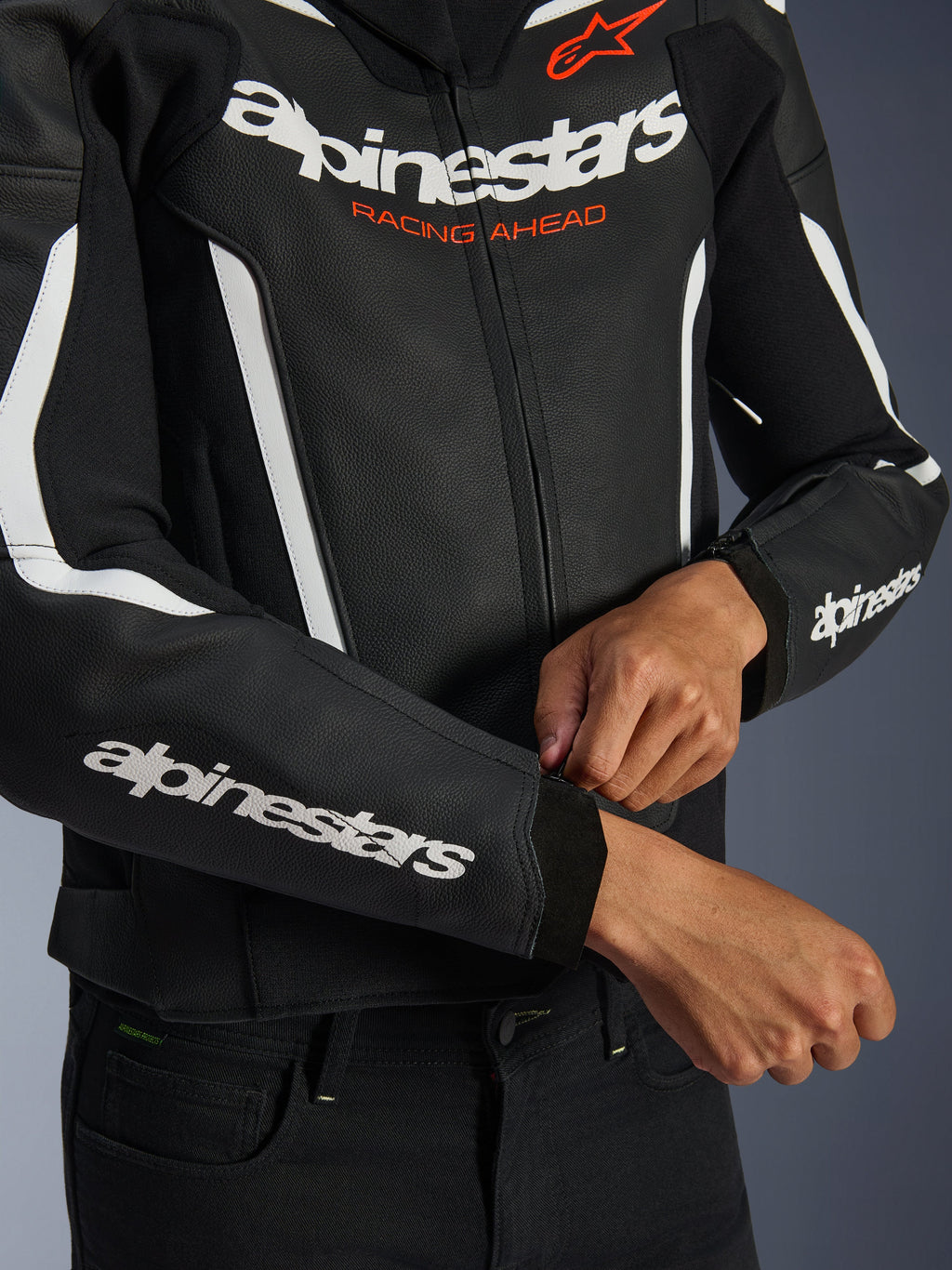 Giacca in pelle Alpinestars GP Force V2, nero e bianco, primo piano del pilota che regola la cerniera del polsino, branding bianco sul petto e sulle maniche con testo 'Racing Ahead' arancione, pelle bovina premium per la guida sportiva in moto