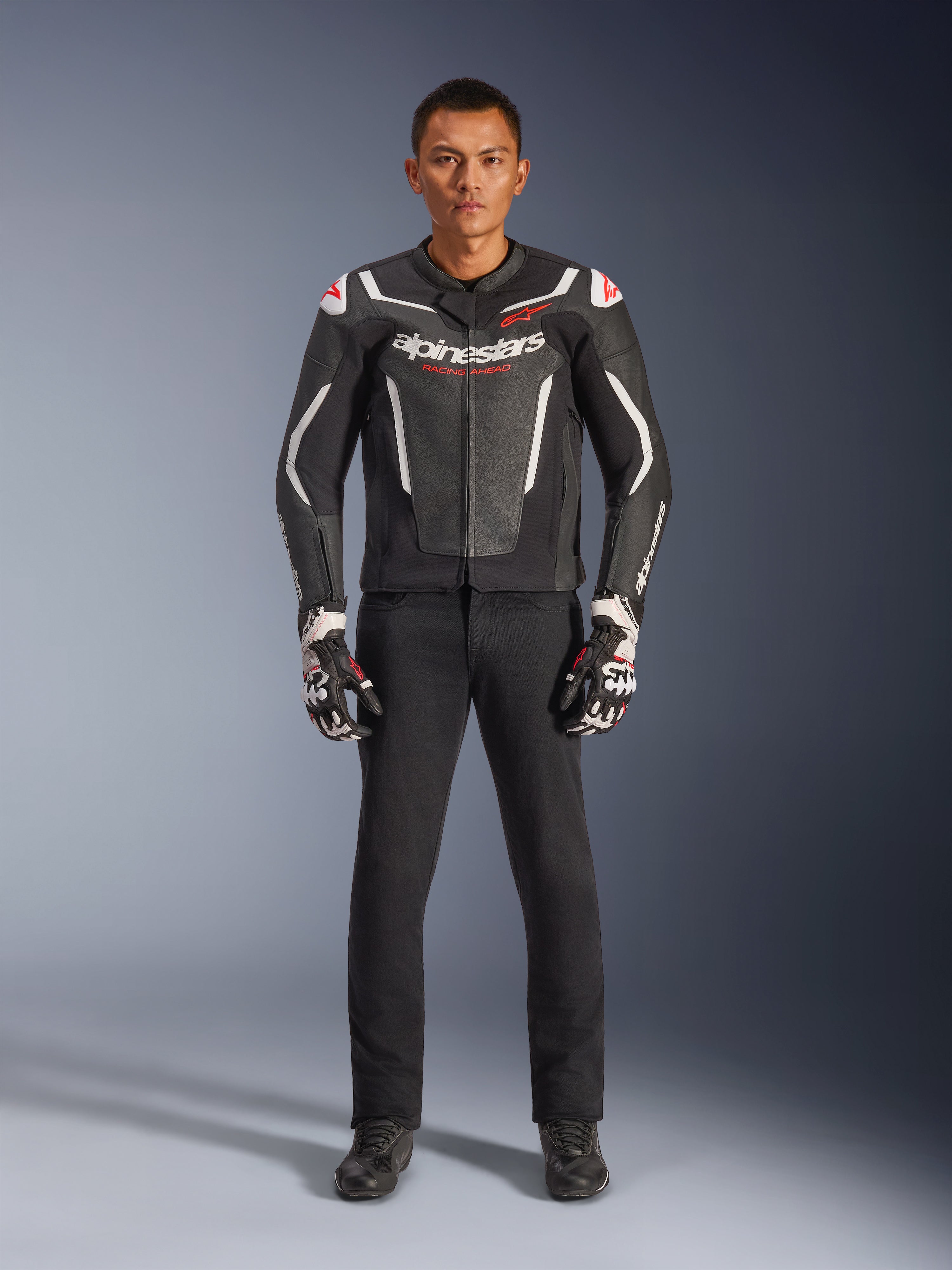 Modello che indossa la giacca in pelle Alpinestars GP Force V2 in nero e bianco, abbinata a pantaloni da moto neri e guanti bianchi e neri, con marchio Alpinestars in evidenza e accenti rossi sulle spalle, abbigliamento professionale per la guida sportiva in moto