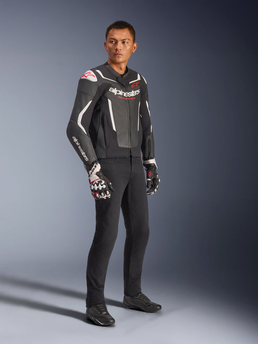 Giacca in pelle Alpinestars GP Force V2, nero e bianco, indossata da un modello con pantaloni da guida neri e guanti tecnici, caratterizzata da loghi Alpinestars rossi, giacca da moto in pelle sportiva per la guida su strada