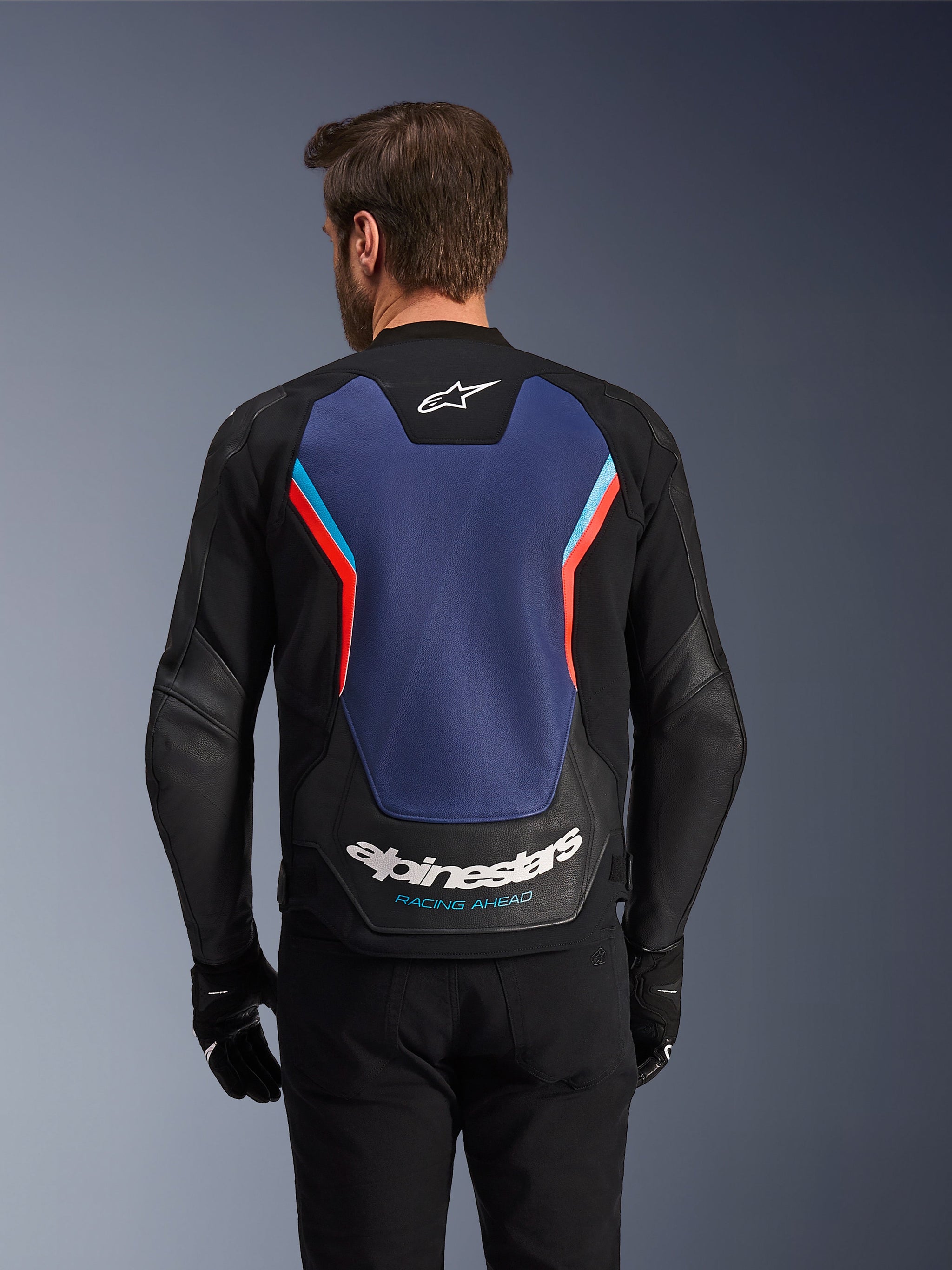 Vista posteriore della giacca in pelle Alpinestars GP Force V2 in nero, blu e rosso fluo, caratterizzata da un ampio pannello posteriore blu con accenti rosso fluorescente e azzurro, logo Alpinestars bianco e scritta 'Racing Ahead' sulla parte bassa della schiena, indossata da un pilota con guanti e pantaloni neri.