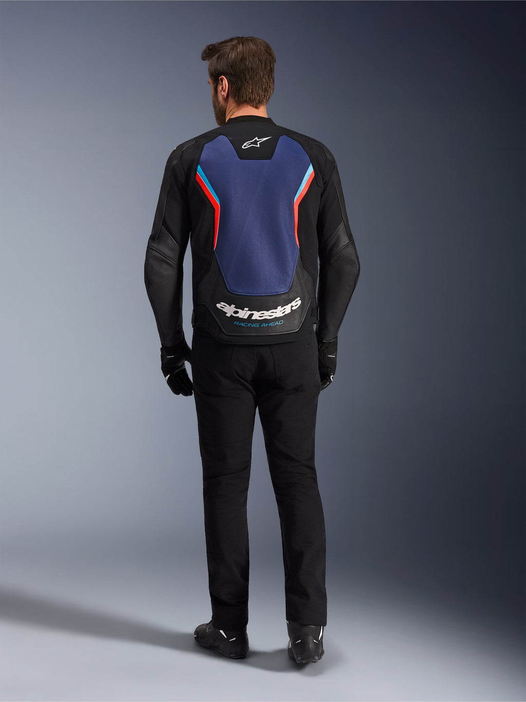 Giacca in pelle Alpinestars GP Force V2, giacca da moto sportiva, Nero Blu Rosso Fluo, vista posteriore su un modello, caratterizzata da un ampio pannello posteriore blu con accenti rossi e azzurri e marchio Alpinestars bianco sulla parte inferiore della schiena, abbinata a pantaloni da moto e guanti neri.