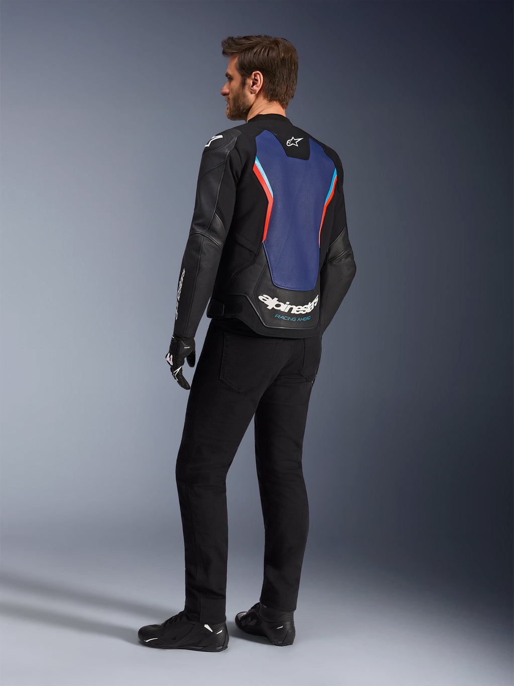 Vista posteriore a tre quarti di un modello che indossa la giacca in pelle Alpinestars GP Force V2 in Black Blue Red Fluo, che mette in risalto il pannello posteriore blu con accenti rosso fluorescente e logo bianco, abbinata a pantaloni neri e guanti da moto.