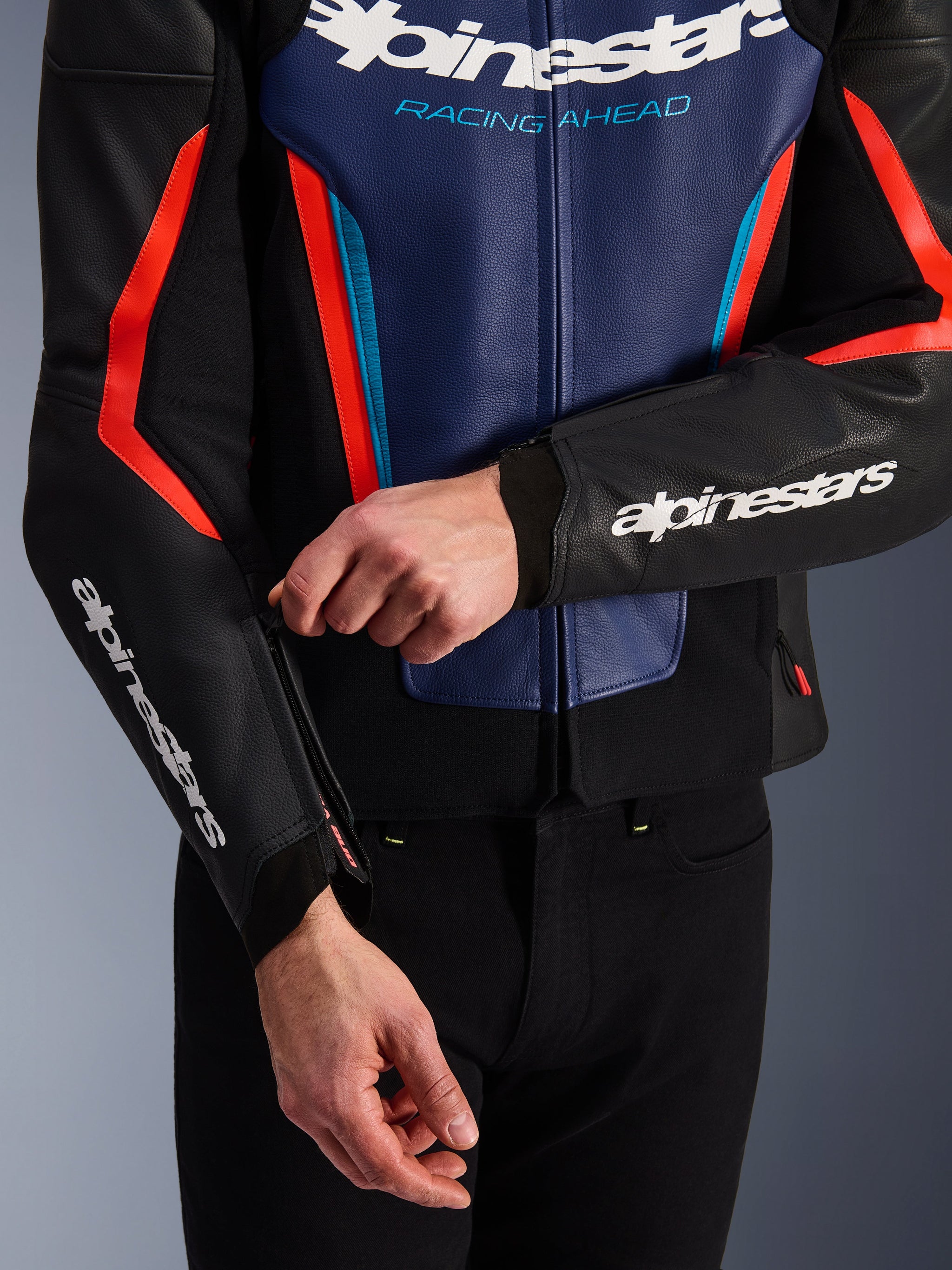 Giacca in pelle Alpinestars GP Force V2 in nero, blu e rosso fluo, vista ravvicinata di una persona che regola la cerniera del polsino, con loghi Alpinestars bianchi su avambraccio e petto, costruzione in pelle bovina testurizzata per guida sportiva