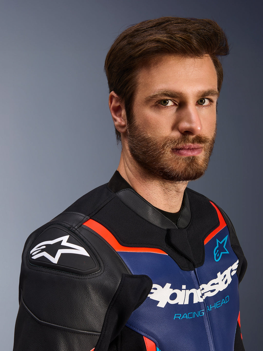 Giacca in pelle Alpinestars GP Force V2, nero, blu e rosso fluo, ritratto ravvicinato di un pilota, con logo Alpinestars bianco sugli scudi DFS neri per le spalle, pannello sul petto in pelle blu con marchio bianco e scritta 'Racing Ahead', accenti rosso fluo, abbigliamento sportivo da moto premium.