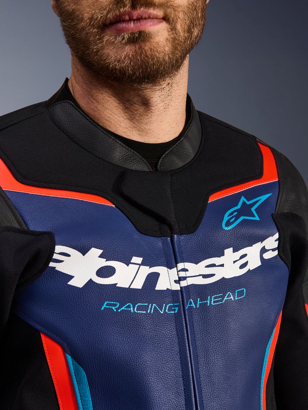 Giacca in pelle Alpinestars GP Force V2, vista dettagliata di petto e colletto, blu, nero e rosso fluo, con logo Alpinestars bianco e scritta 'RACING AHEAD' azzurra, texture in pelle premium con accenti fluorescenti