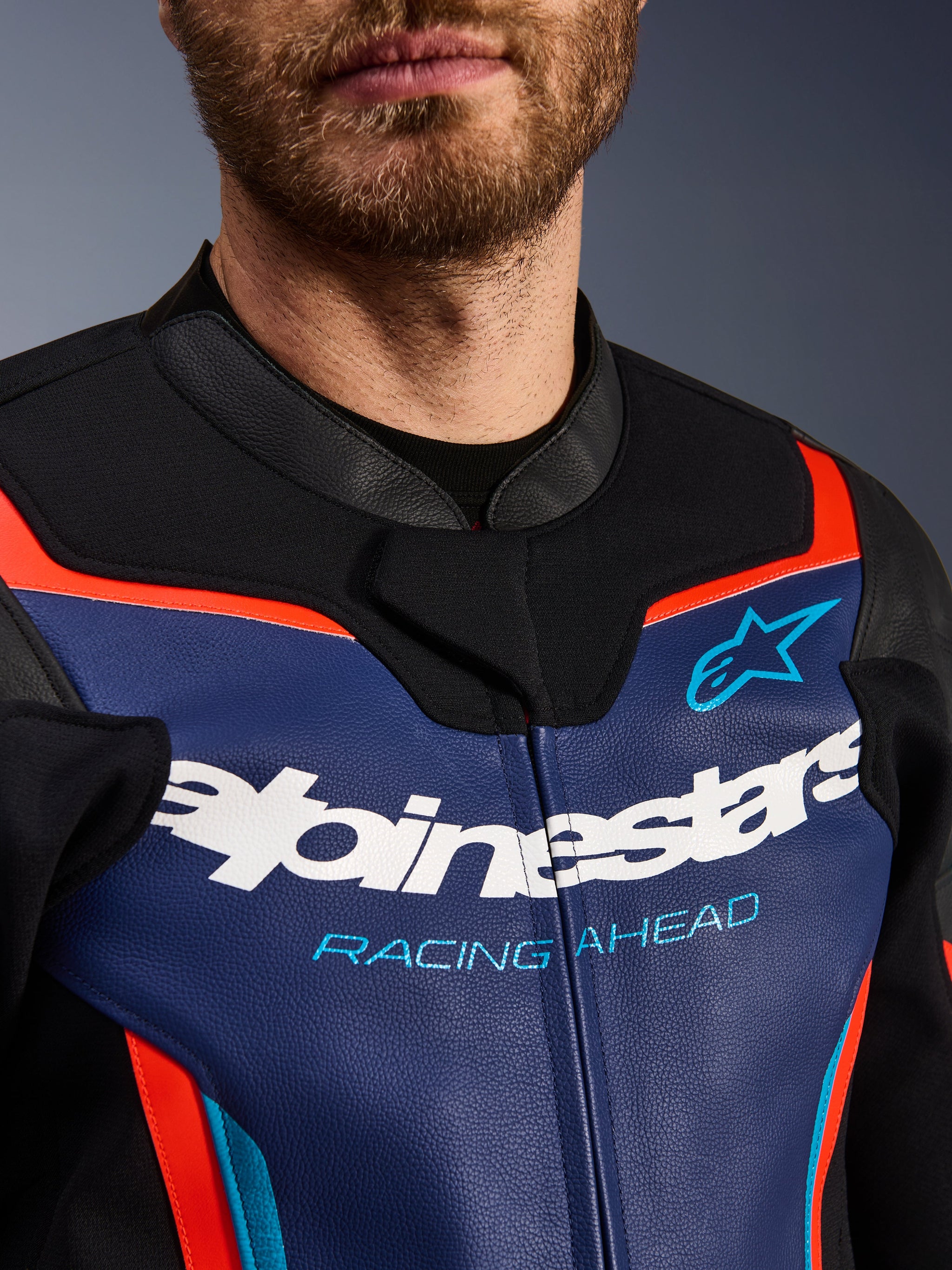 Giacca in pelle Alpinestars GP Force V2, vista dettagliata di petto e colletto, blu, nero e rosso fluo, con logo Alpinestars bianco e scritta 'RACING AHEAD' azzurra, texture in pelle premium con accenti fluorescenti