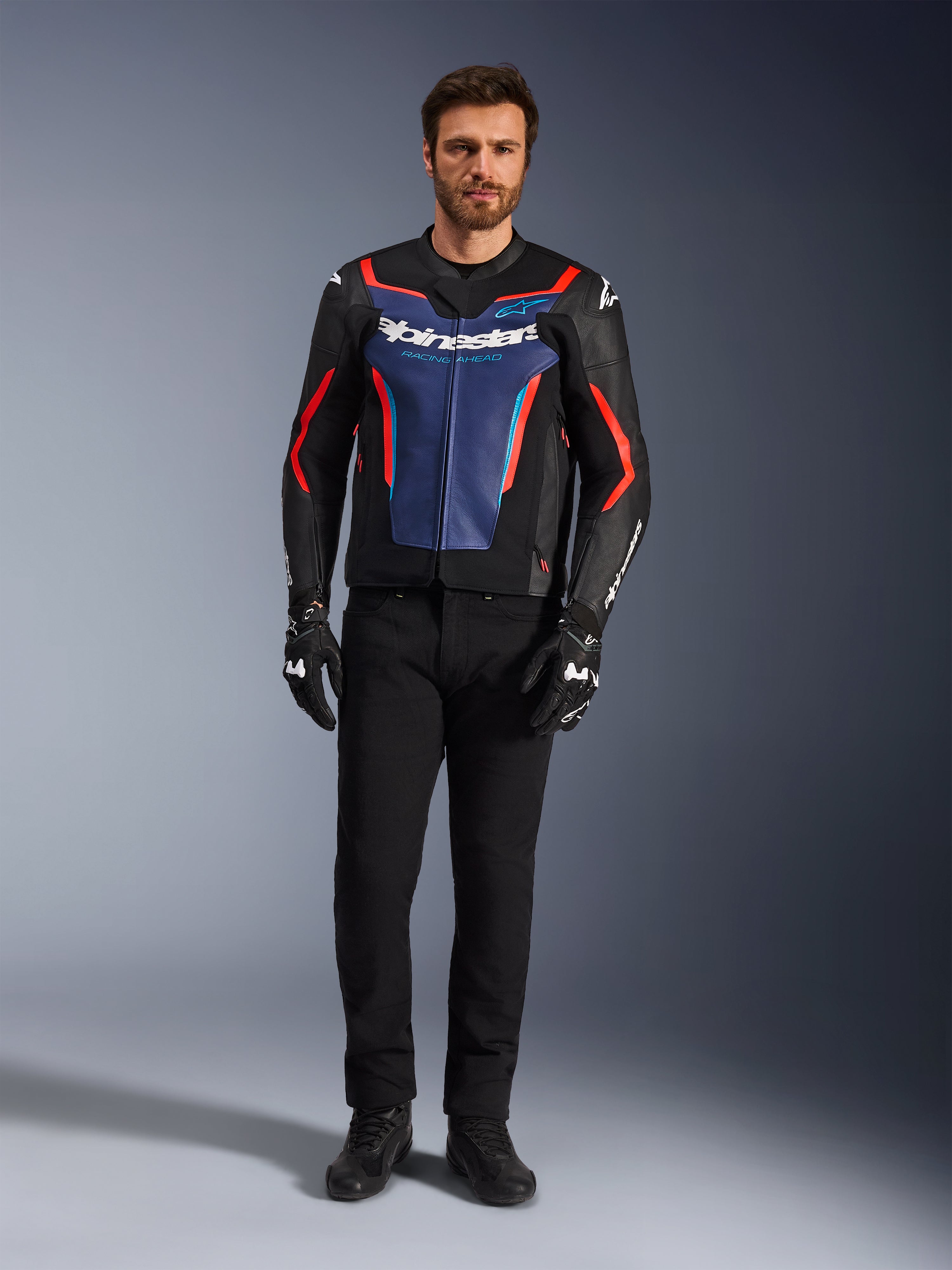 Giacca in pelle Alpinestars GP Force V2, Nero Blu e Rosso Fluo, indossata da un modello uomo con pantaloni neri e guanti da moto, giacca da moto sportiva in pelle bovina dal design d'ispirazione racing, vista frontale