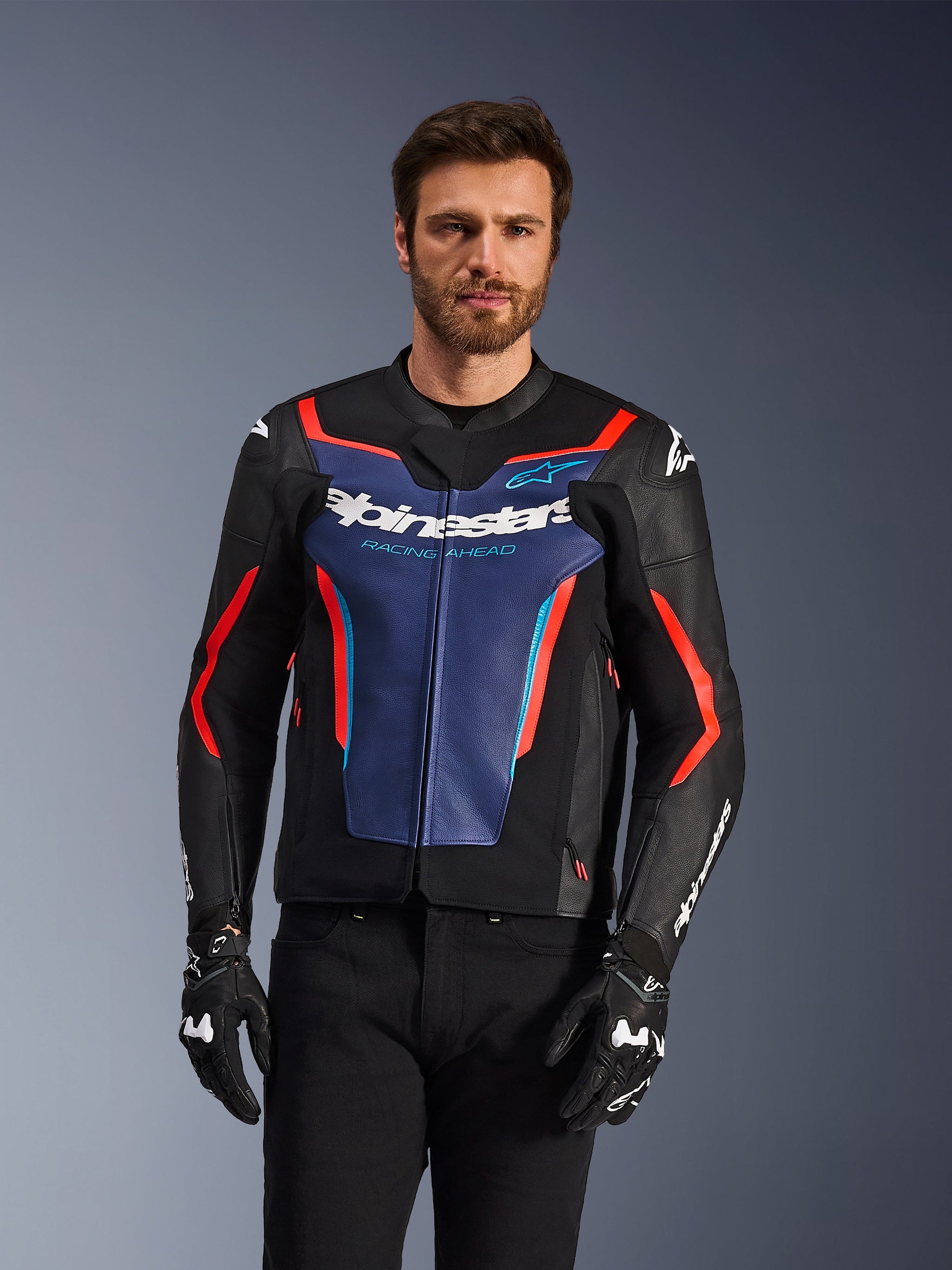 Giacca in pelle Alpinestars GP Force V2, nero, blu e rosso fluo, indossata da un modello con guanti e pantaloni neri, caratterizzata da un pannello blu sul petto con scritta 'Alpinestars Racing Ahead', dettagli rossi e scudi esterni sulle spalle, abbigliamento protettivo da moto sportiva
