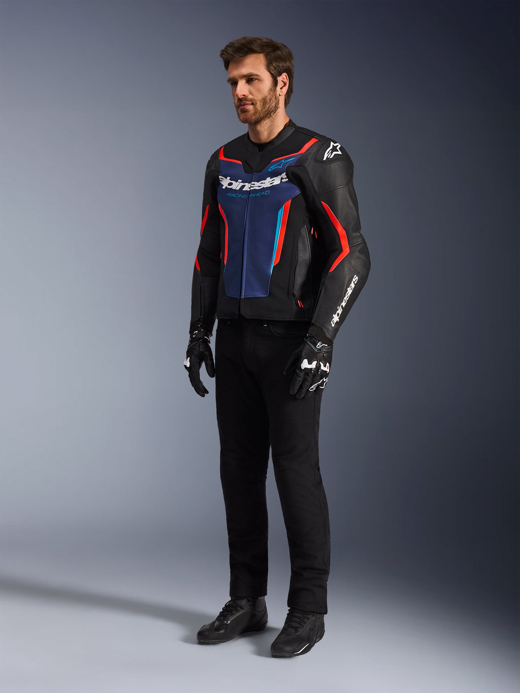 Modello che indossa la giacca in pelle Alpinestars GP Force V2 in Black Blue Red Fluo, abbinata a pantaloni tecnici neri, guanti e stivali neri, abbigliamento professionale da motociclismo per guida sportiva