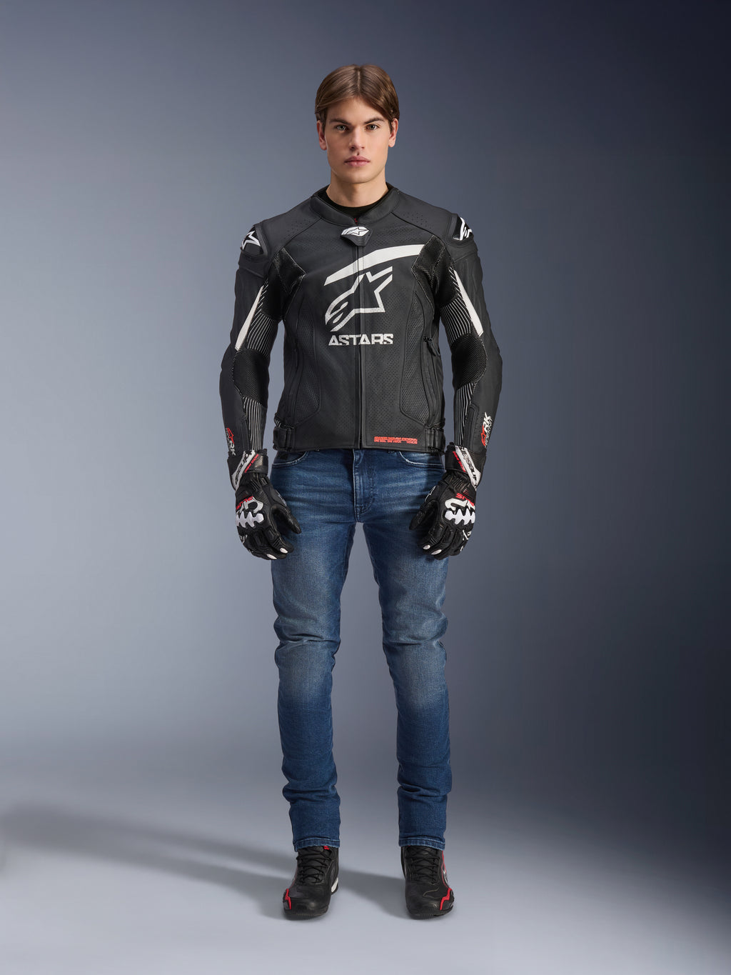 Modello che indossa la giacca in pelle Alpinestars GP Plus R V4 Rideknit in nero e bianco, abbinata a jeans da moto blu, guanti da moto neri e scarpe da guida nere, con un grande logo Astars bianco sul petto, pannelli in pelle traforata e strisce racing bianche sulle maniche, vista frontale a figura intera