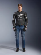 Modello che indossa la giacca in pelle Alpinestars GP Plus R V4 Rideknit in nero e bianco, abbinata a jeans da moto blu, guanti da moto neri e scarpe da guida nere, con un grande logo Astars bianco sul petto, pannelli in pelle traforata e strisce racing bianche sulle maniche, vista frontale a figura intera